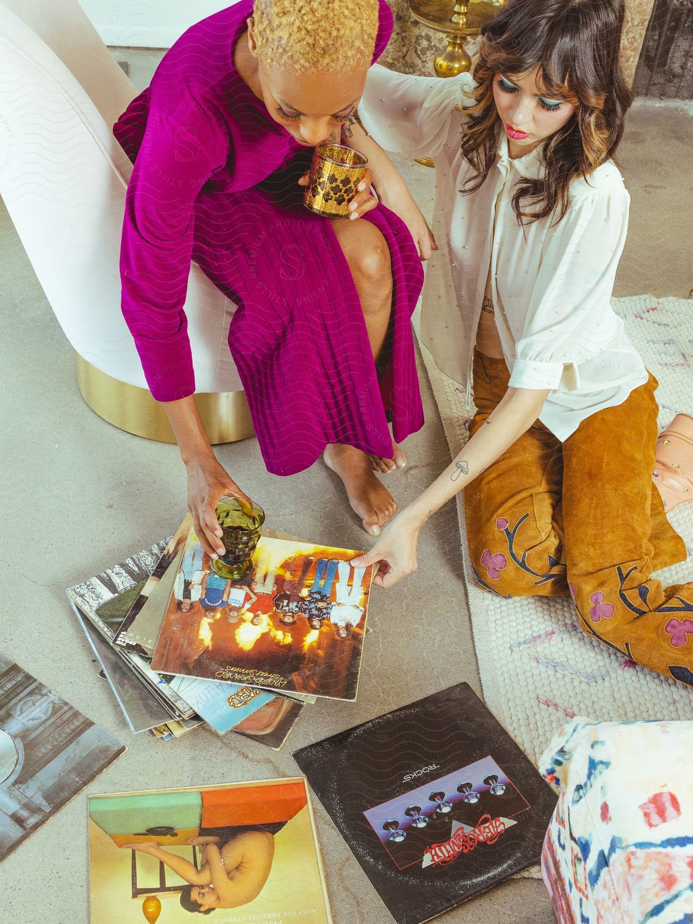 A woman sitting in a white armchair and another sitting on a rug on the floor and both are holding a record preventing what is on the floor and one of them placing a glass on top of the record cover