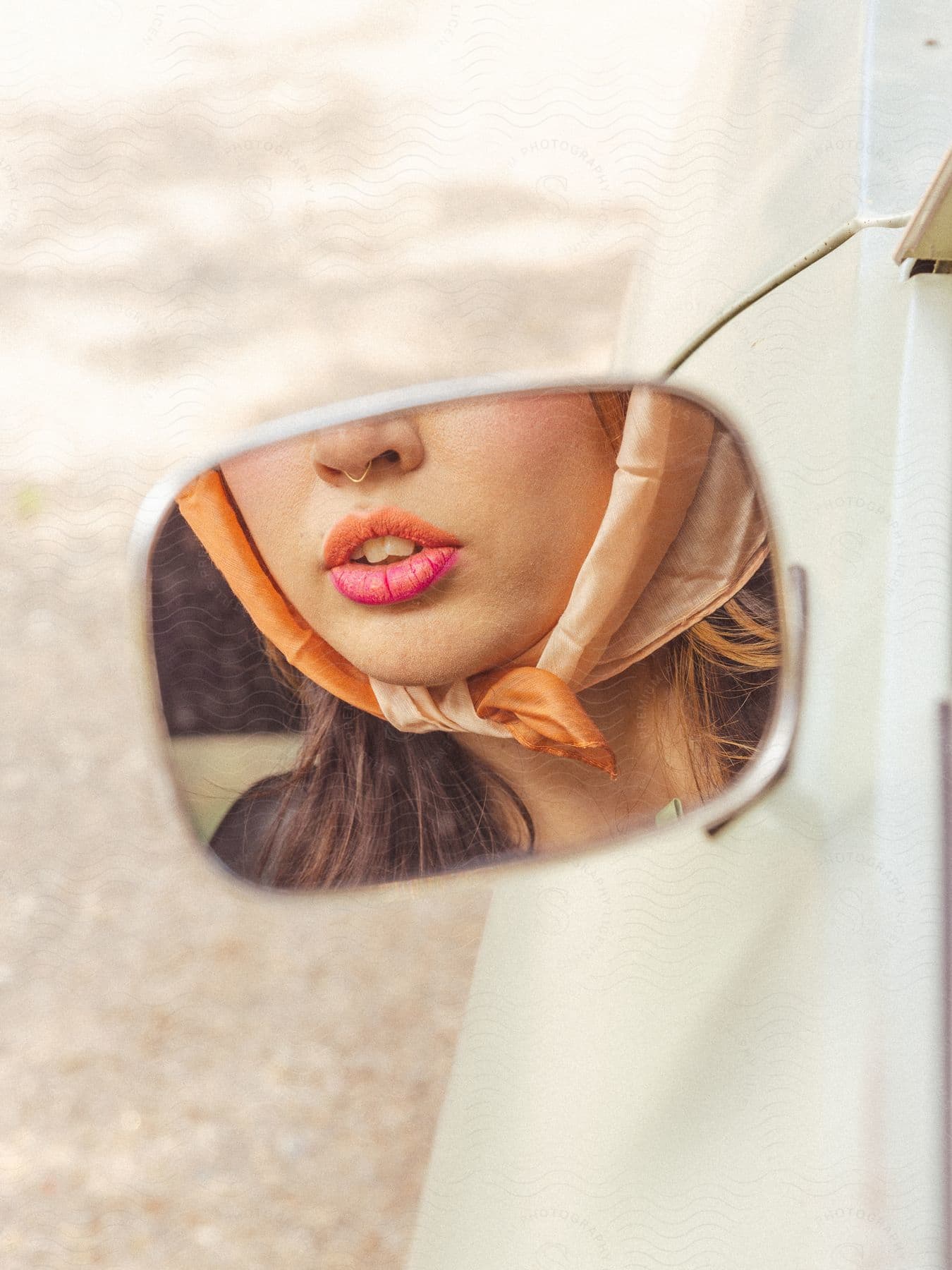A woman's face reflected in a car mirror with her lips open and her nose pierced and a cloth on her chin