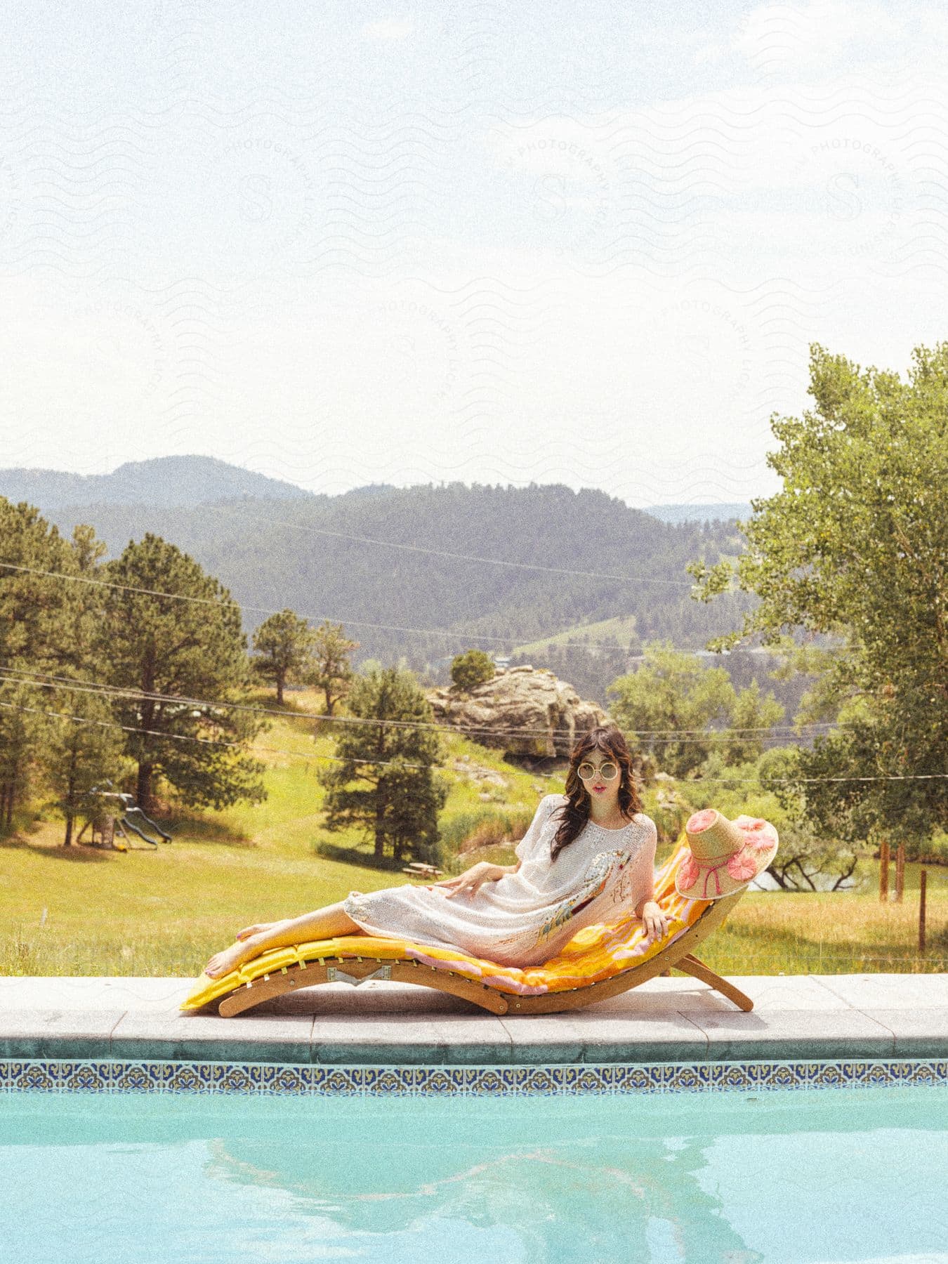 Woman posing on a poolside deck chair with a forested hillside in the background.