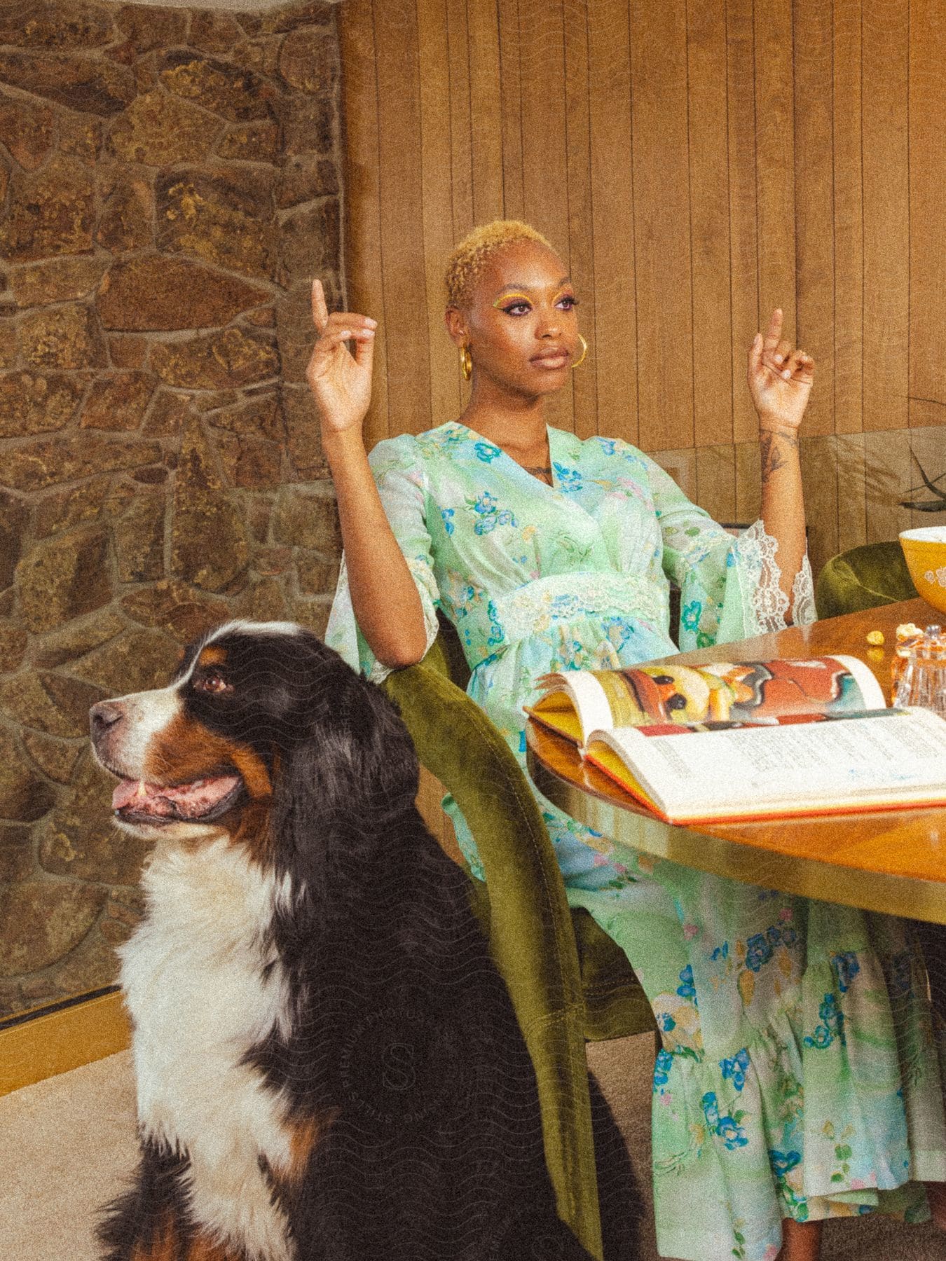 Woman in a green flowery dress sitting in an armchair in front of the table with a dog by her side