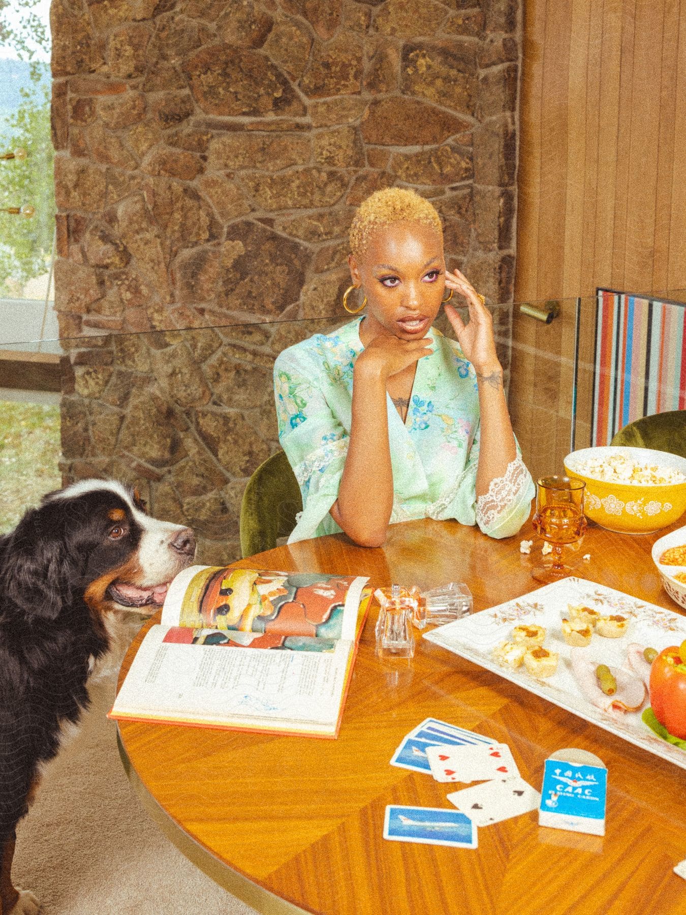 Woman seated at a table with a dog, snacks, playing cards, and a magazine, in a room with stone and wood paneling.