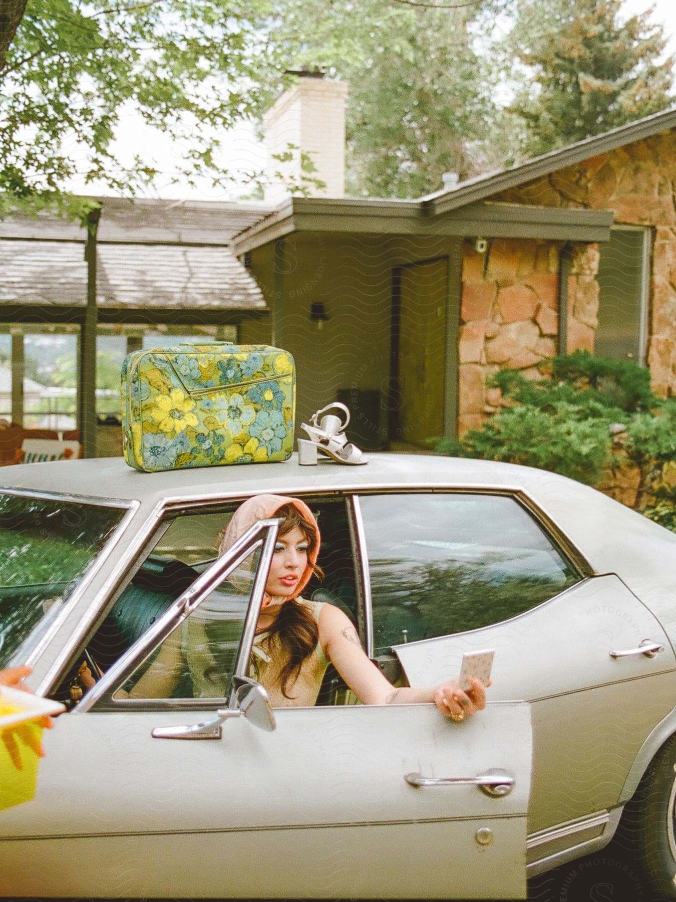 A woman with a hood is getting out of an old car and there is a green bag and shoes on top of the car