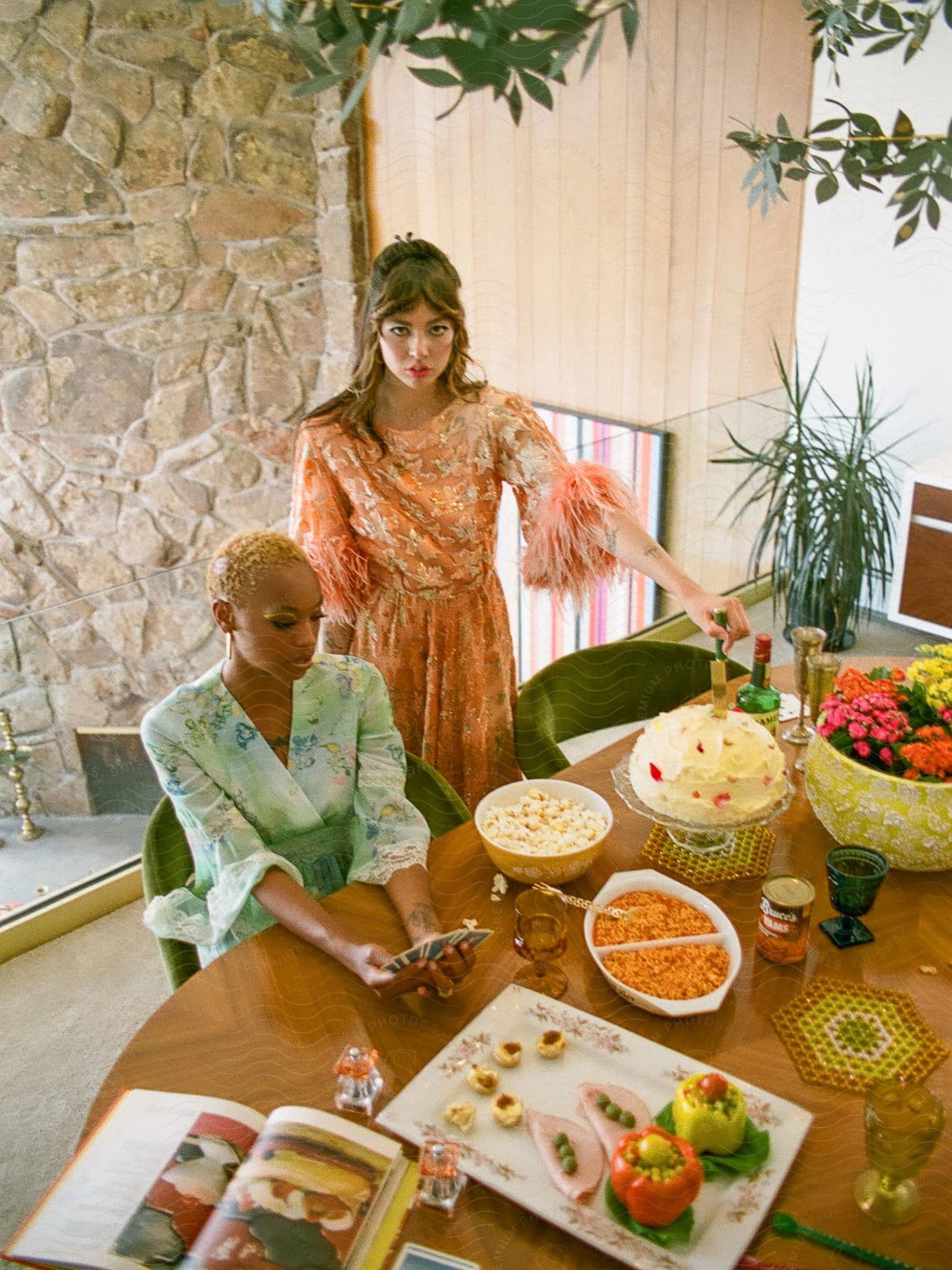 a woman with short hair and a light blue dress sits at a table with meals, holding cards, next to a woman wearing a pink dress standing and looking up