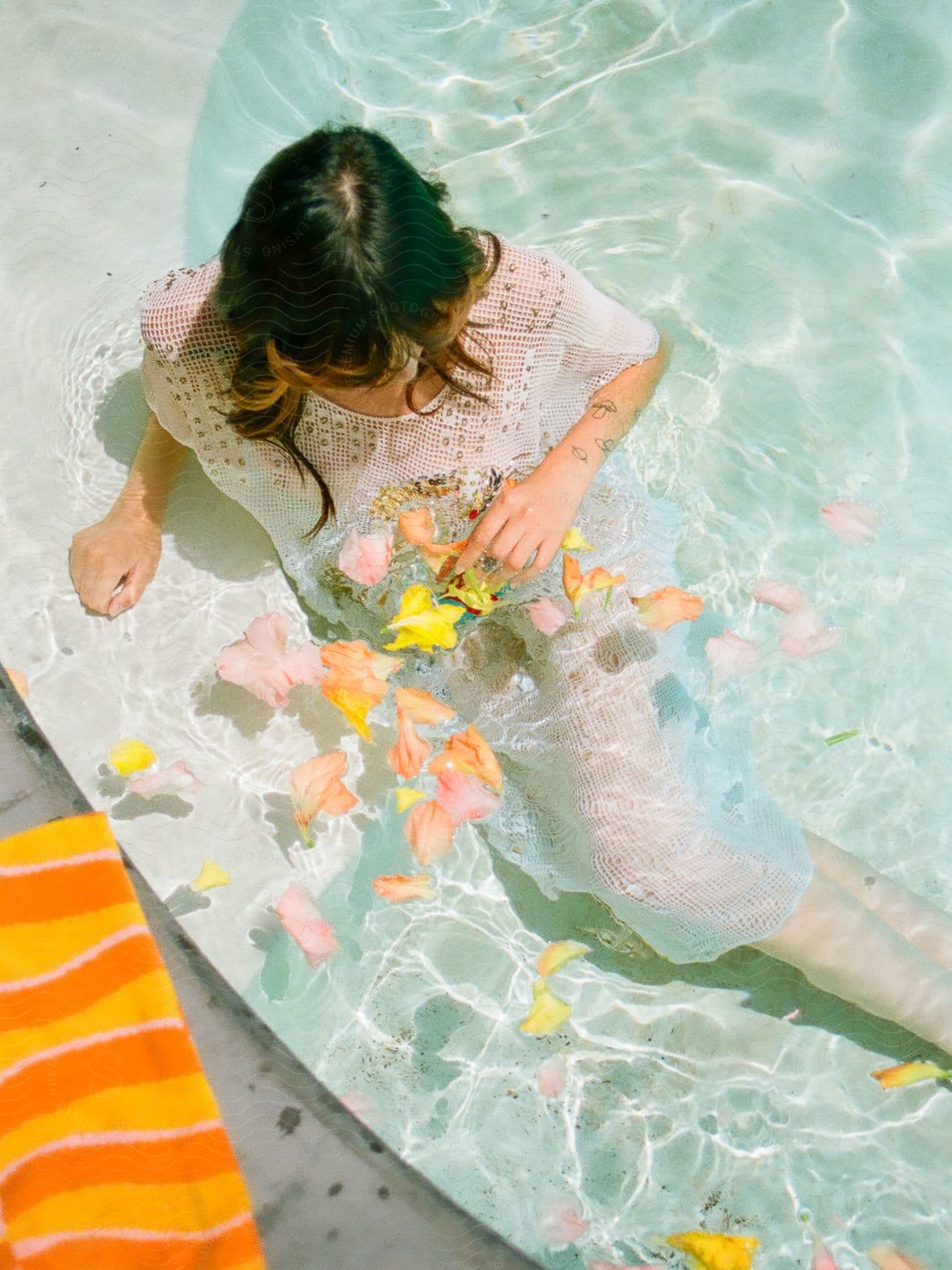 On a sunny day, a woman sits on the poolside surrounded by scattered flowers.