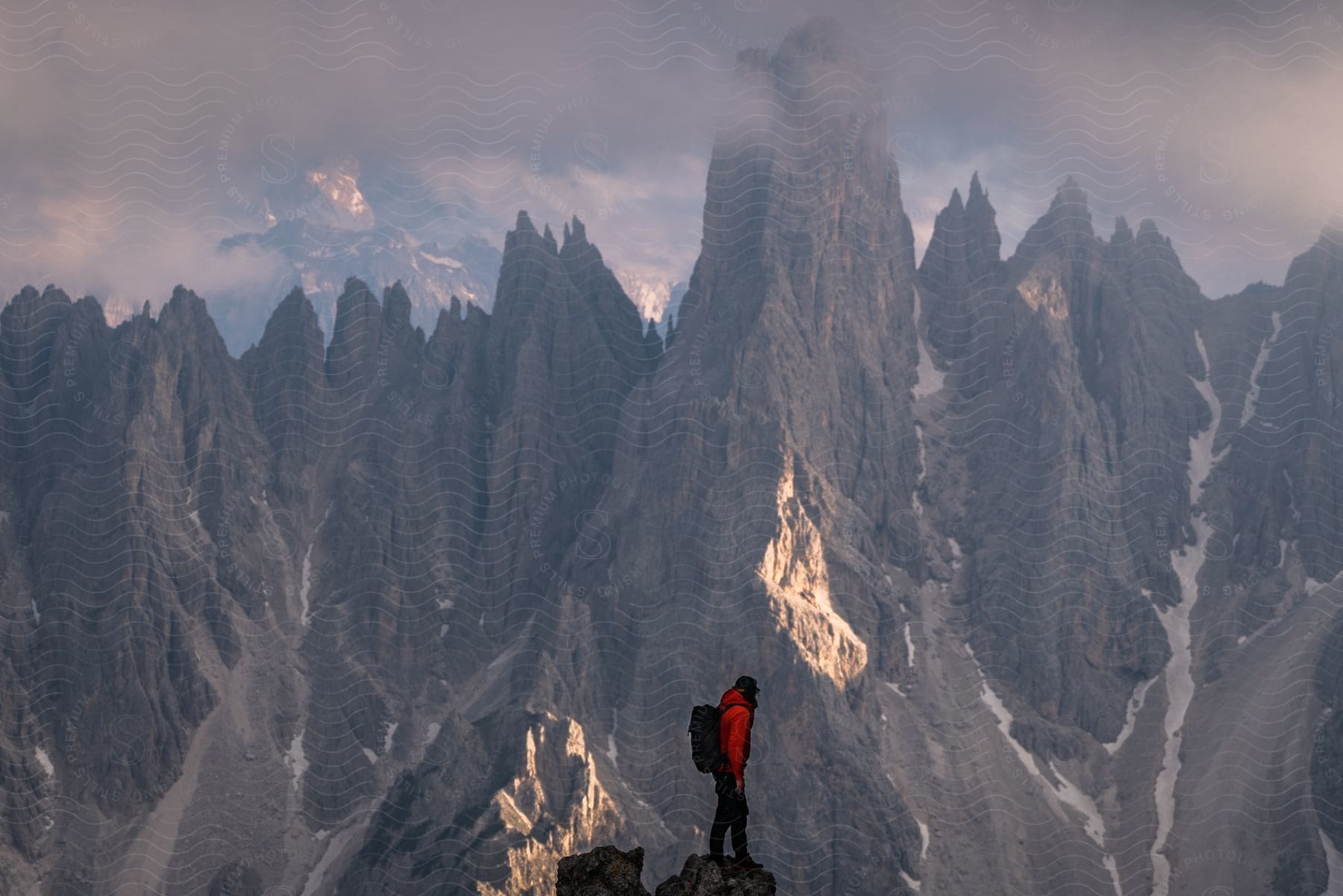 Stock photo of hiker stands on rock formations looking at rough terrain mountains with peaks high in a cloudy sky