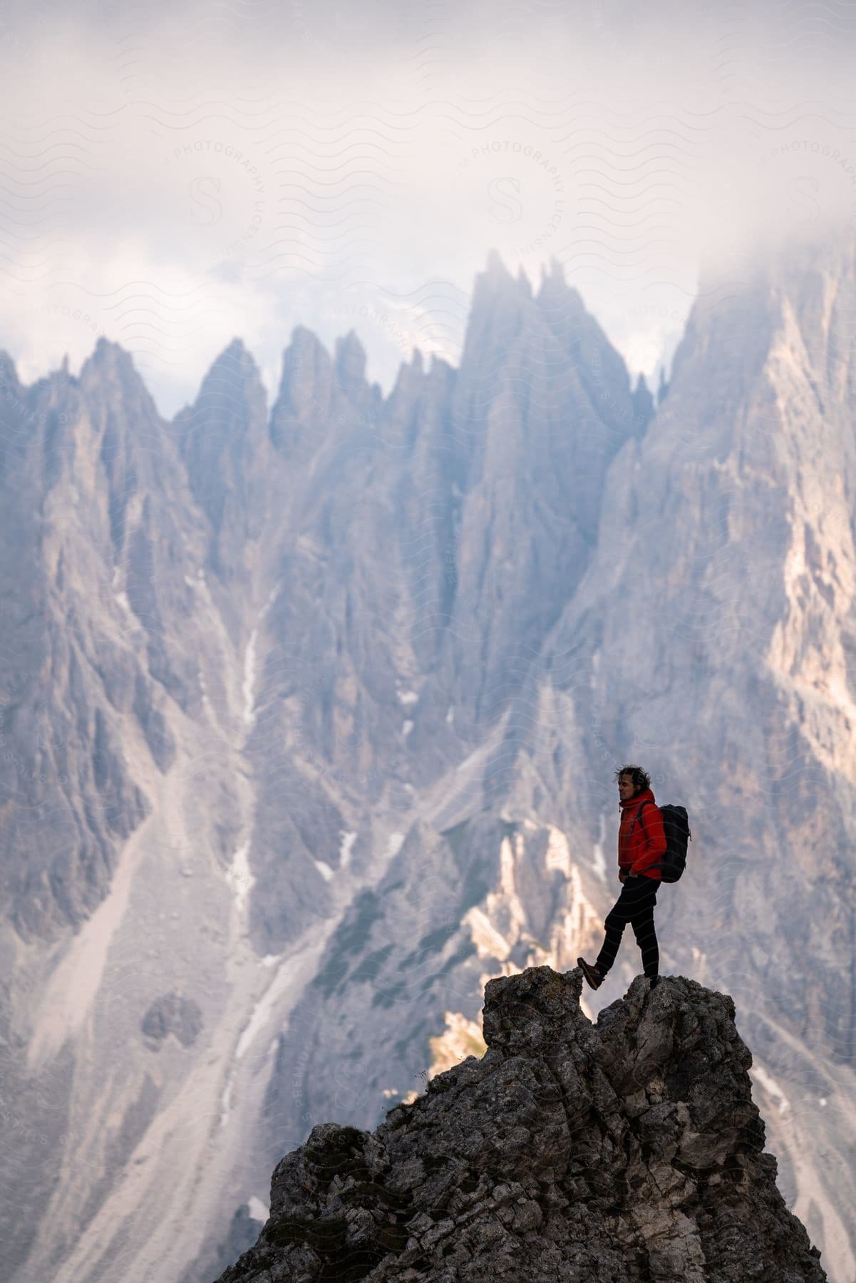 Person standing on a rocky peak with jagged mountain range in the background, wearing a red jacket and carrying a backpack.