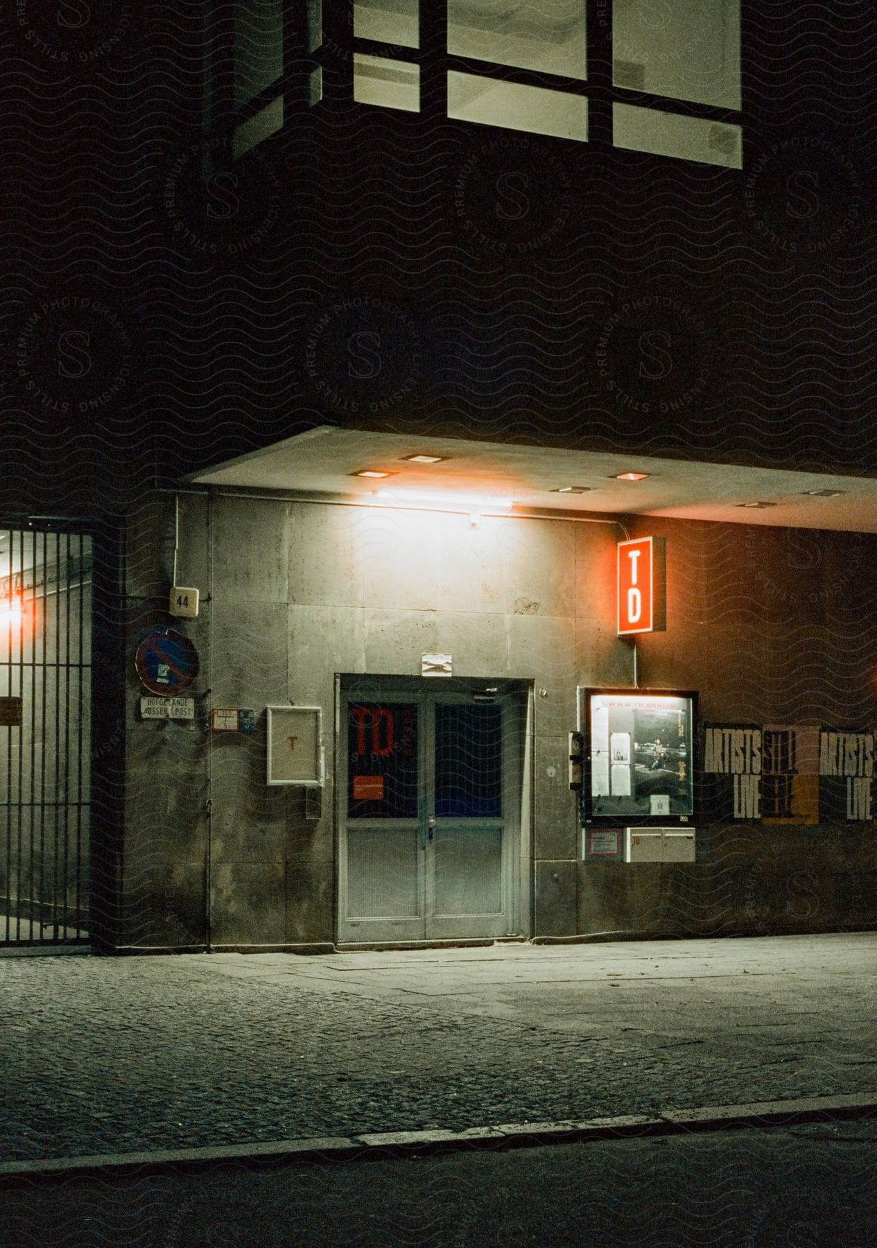 At night, the entrance door to a building is closed, with light shining from the ceiling and through the window above, illuminating the interior.