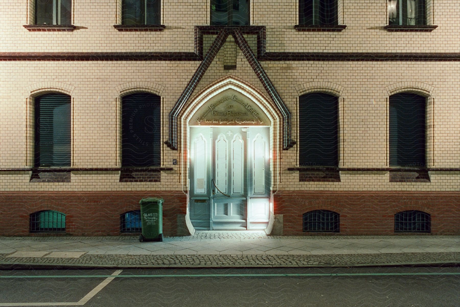 The illuminated entrance of a brick building at night with a green trash can next to it.