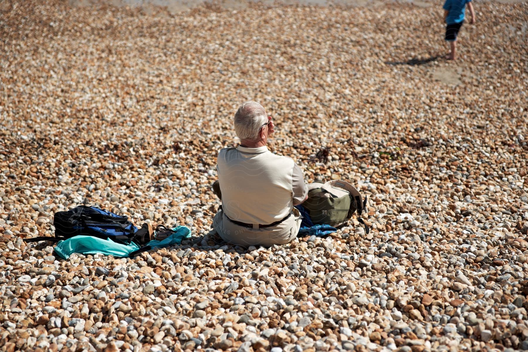 An elderly man relaxes on a rocky shore, his backpack and bag nearby, as a child in blue bursts into view.