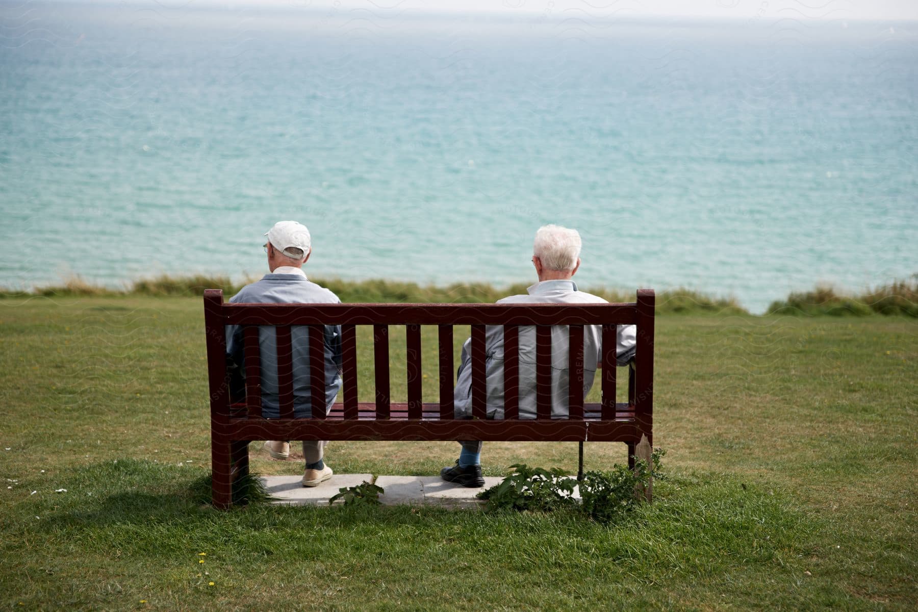 Two elderly men sit on a bench near the sea on a coastal island covered in grass during the day. One of them wears a baseball hat.