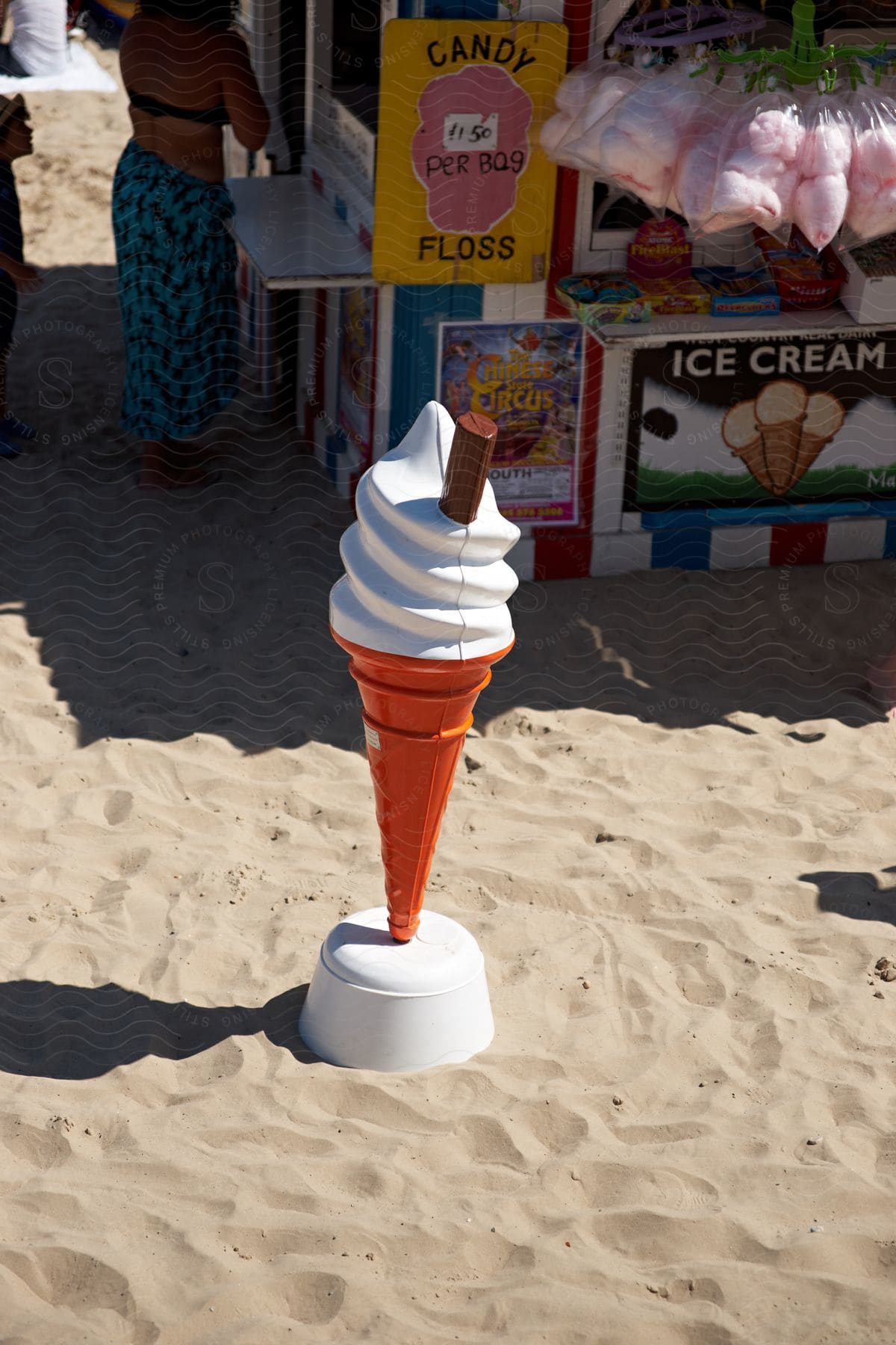 A large model of an ice cream cone with a chocolate bar placed on the sand near a candy and ice cream stand.