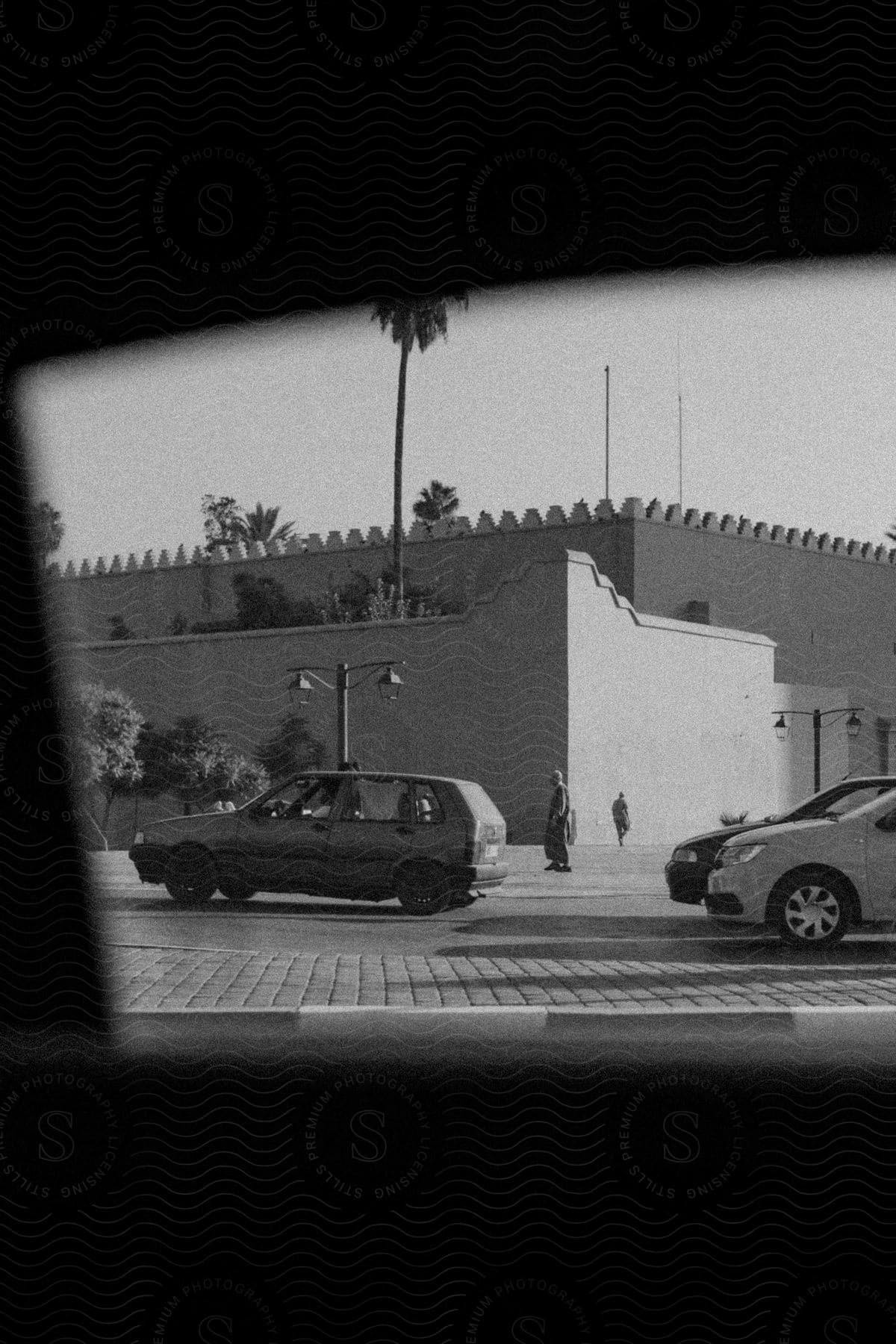 Street with cars and pedestrian, framed by window silhouette, palm trees and building with battlements.