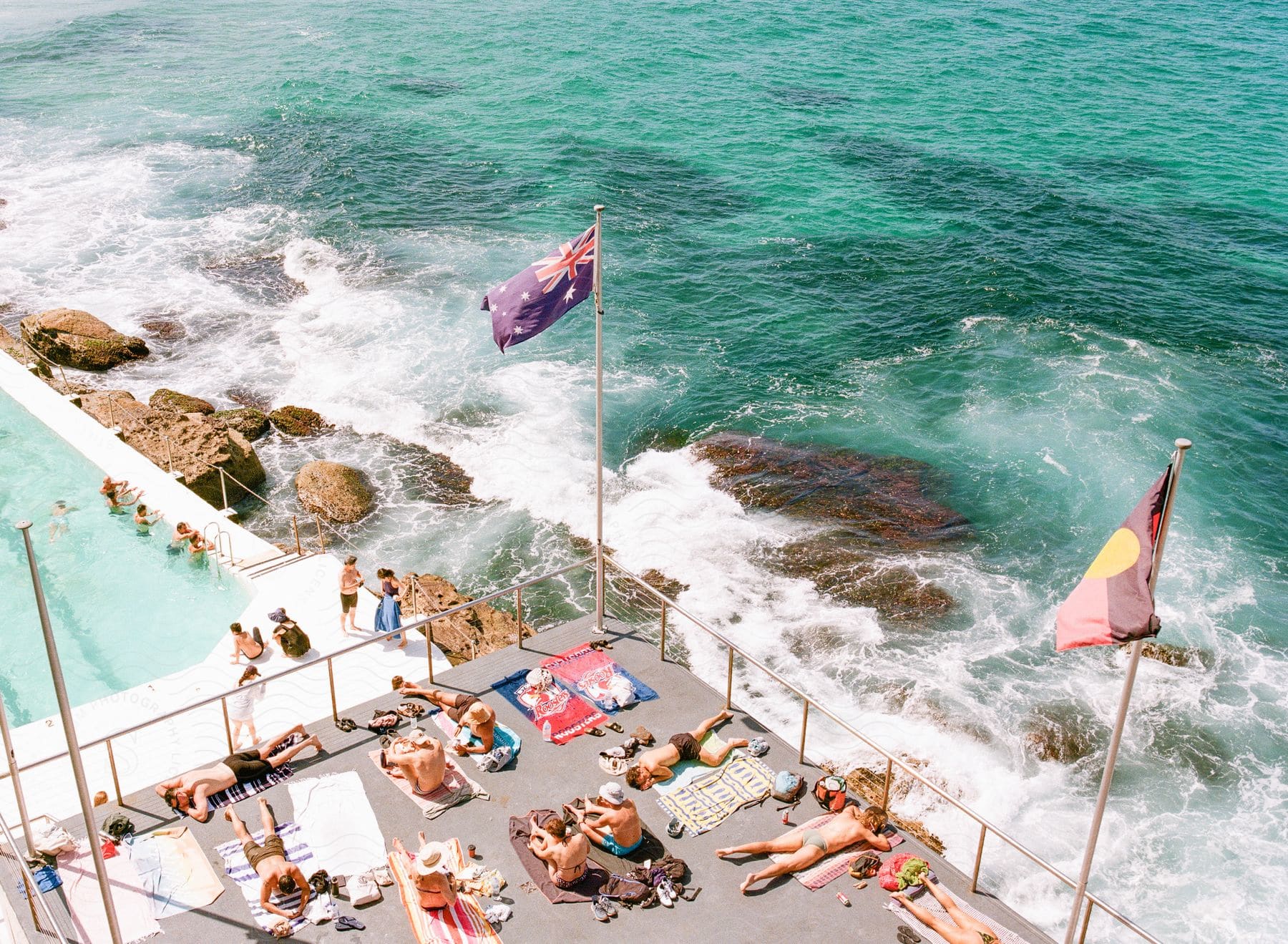 People in the pool and sunbathing on the deck of the Bondi Icebergs Swimming Club as ocean waves splash against the rocks