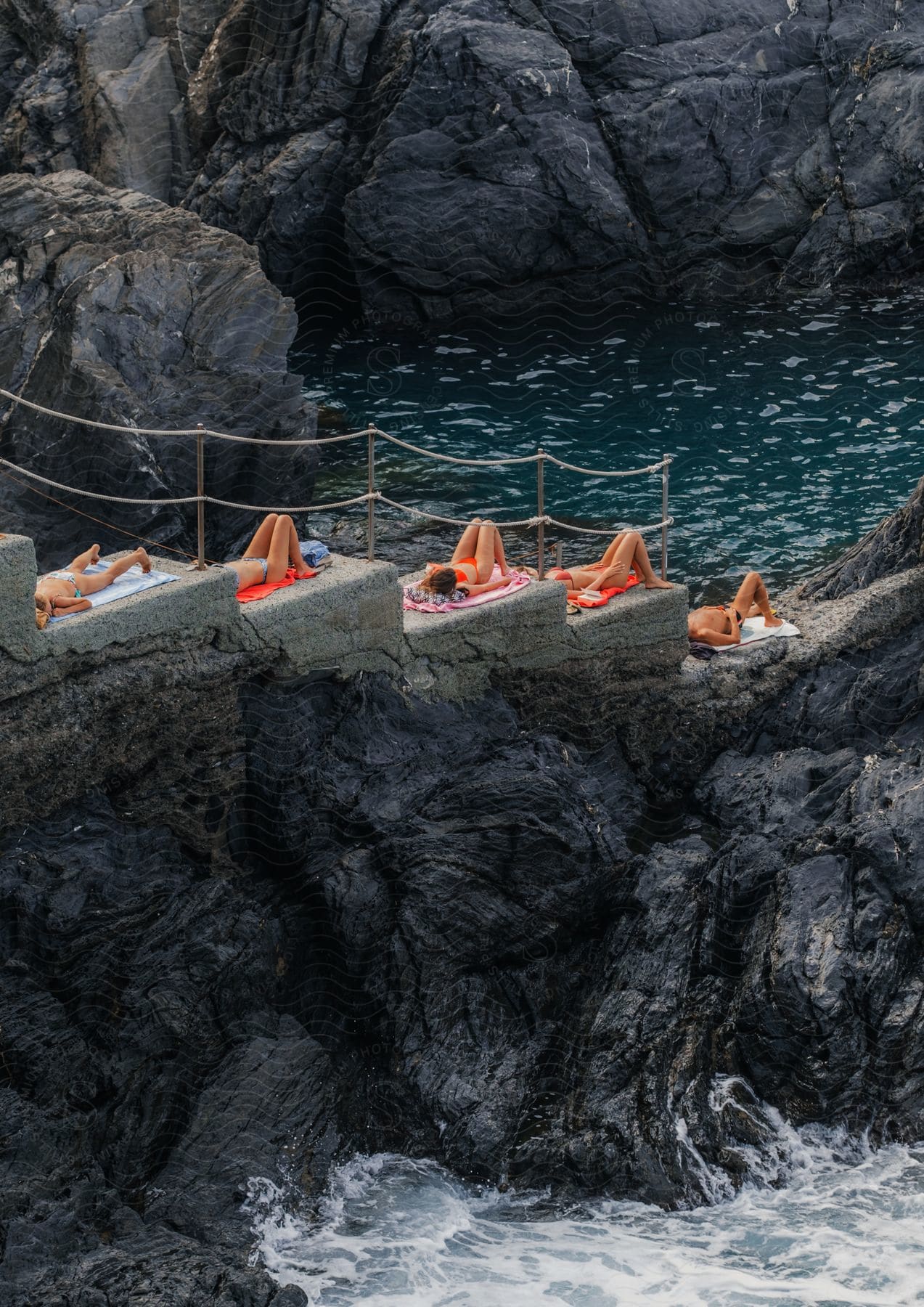 Five adults sunbathe on concrete steps on a rocky ocean coast.