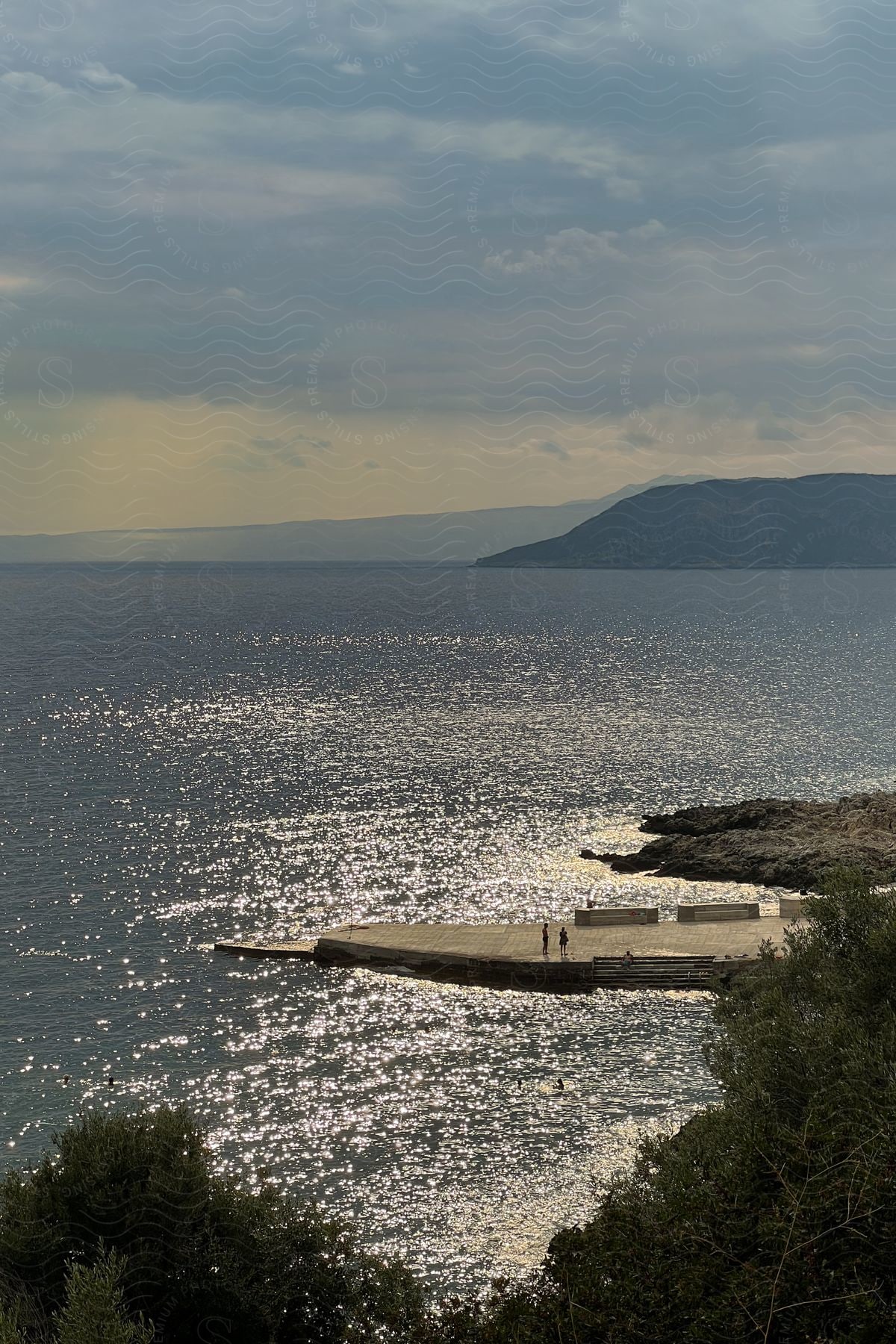 A serene view of a concrete pier stretching out into a sparkling sea, with mountains in the distance under a cloudy sky. People are standing on the edge of the pier, enjoying the view.