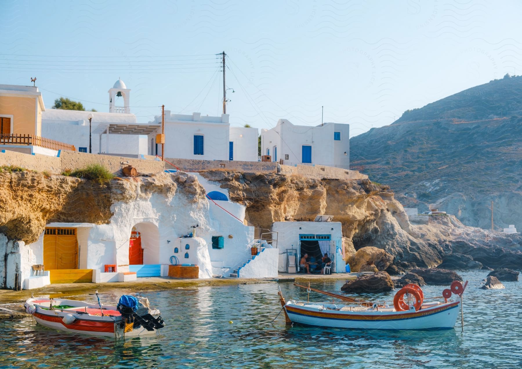 Buildings overlooking the water and boats off the coast as people sit near the water with a mountain in the distance