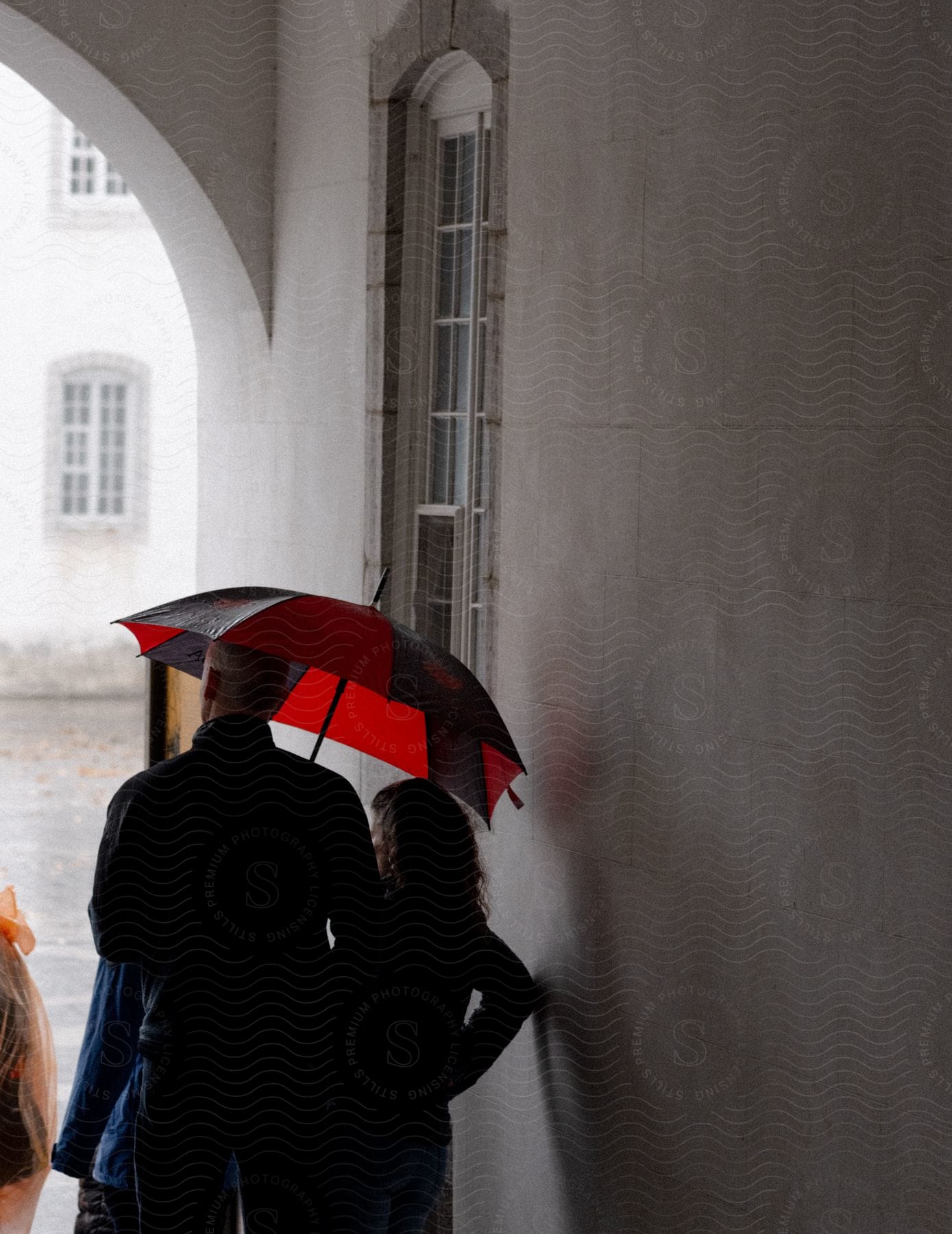 Two people stand under a red umbrella, looking out from an arched corridor during the rainy day.