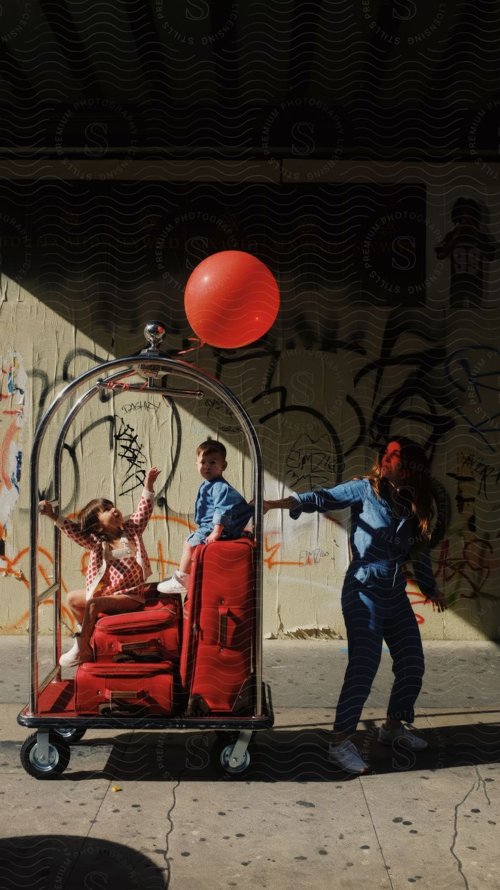 Mother in denim clothes pulling a cart with red suitcases and her two children sitting with a red balloon tied in the environment of a viaduct through the city with graffiti on the walls.