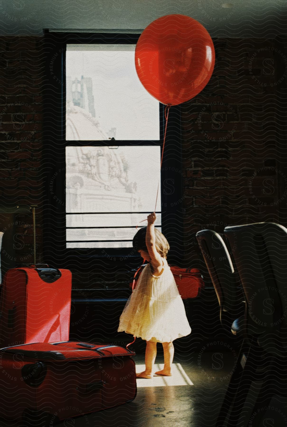 Girl in a white dress holding a red balloon next to red suitcases in a room with sunlight reflections.