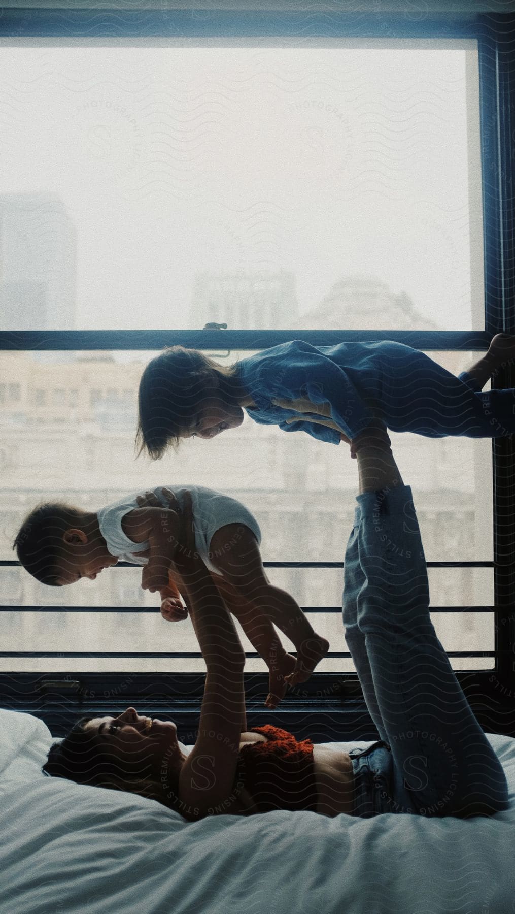 Woman is lying on her bed exercising with her children balancing one on her feet in the air as she holds up another boy with her arms