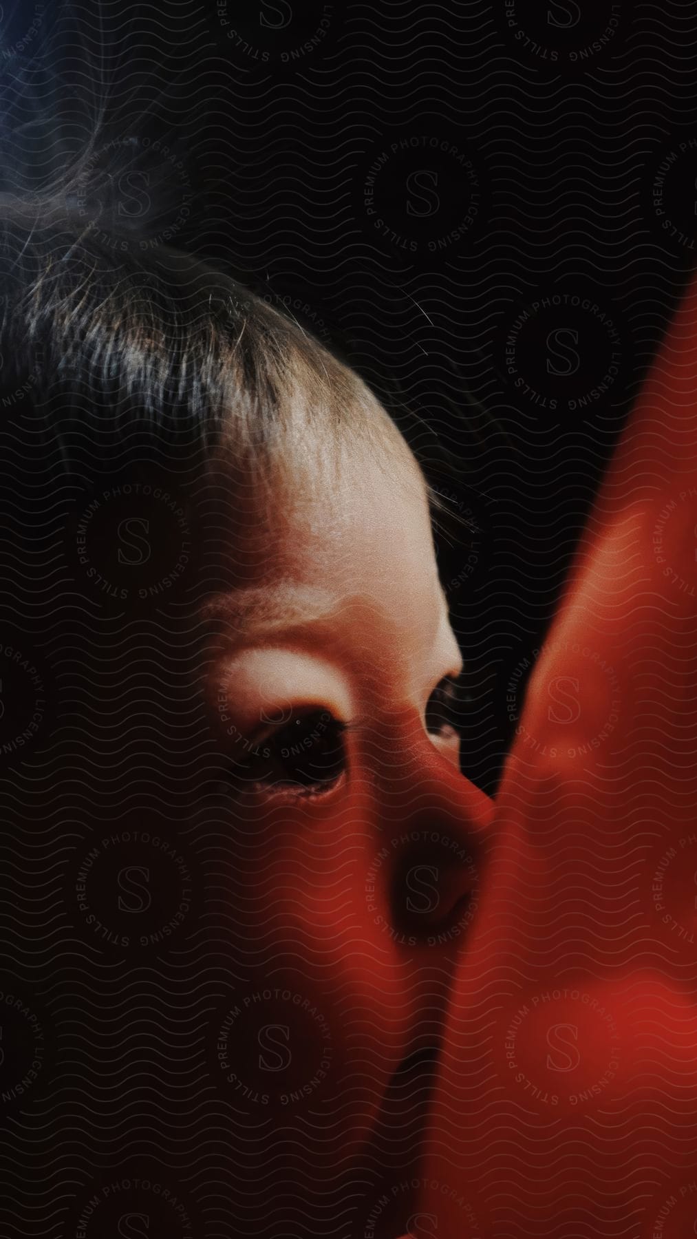 Close-up of a child's side face kissing a red balloon against a black background.