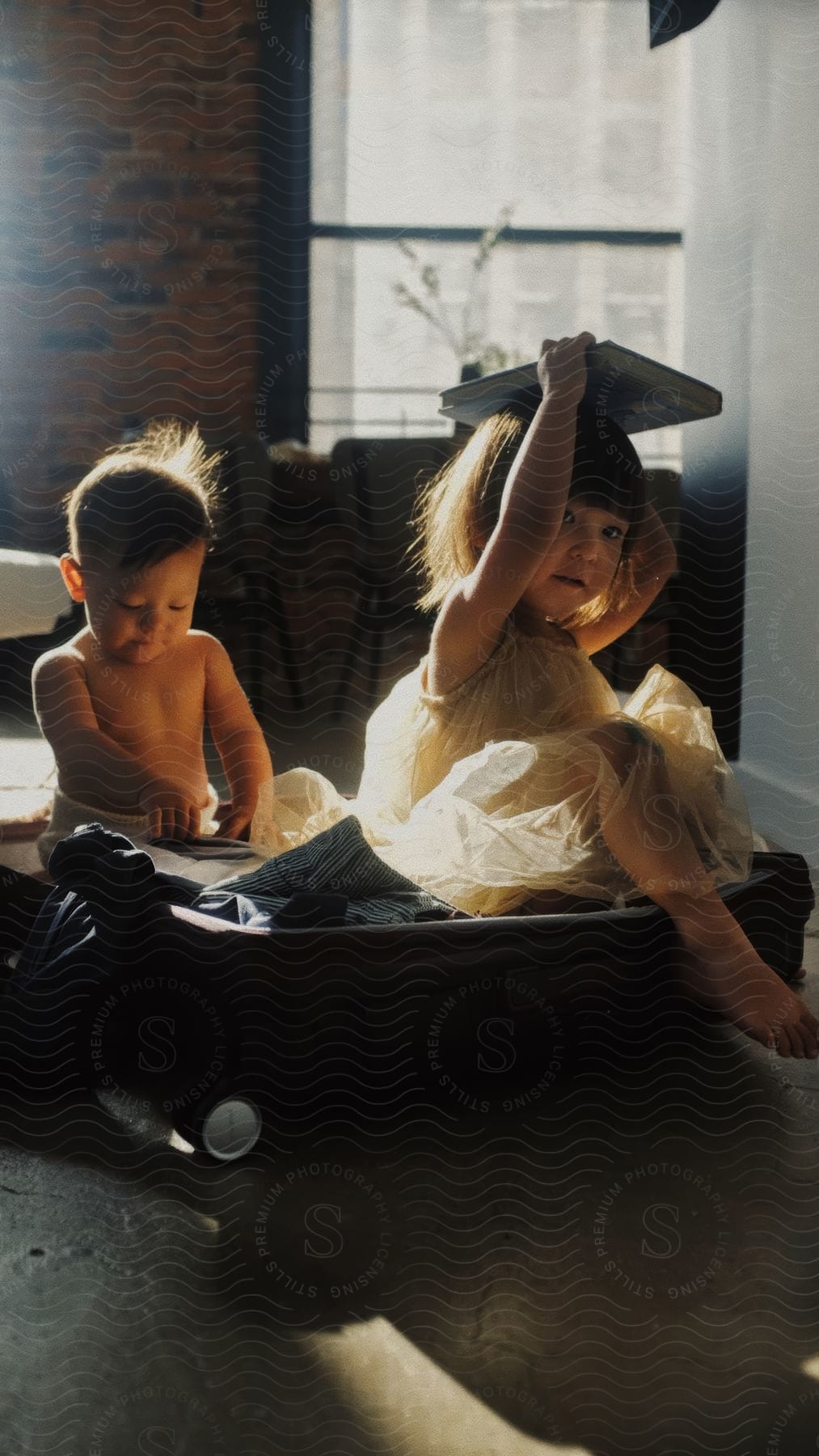 Two children are playing with a suitcase, illuminated by natural light from a nearby window.