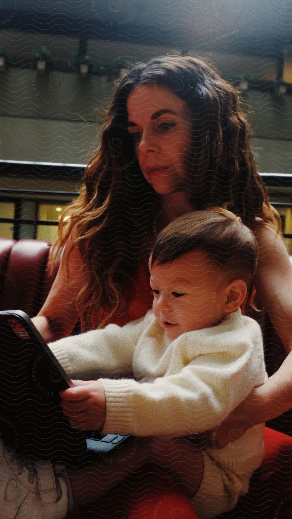 Mother sitting with her baby on her lap on a red upholstered bench with a laptop in use.