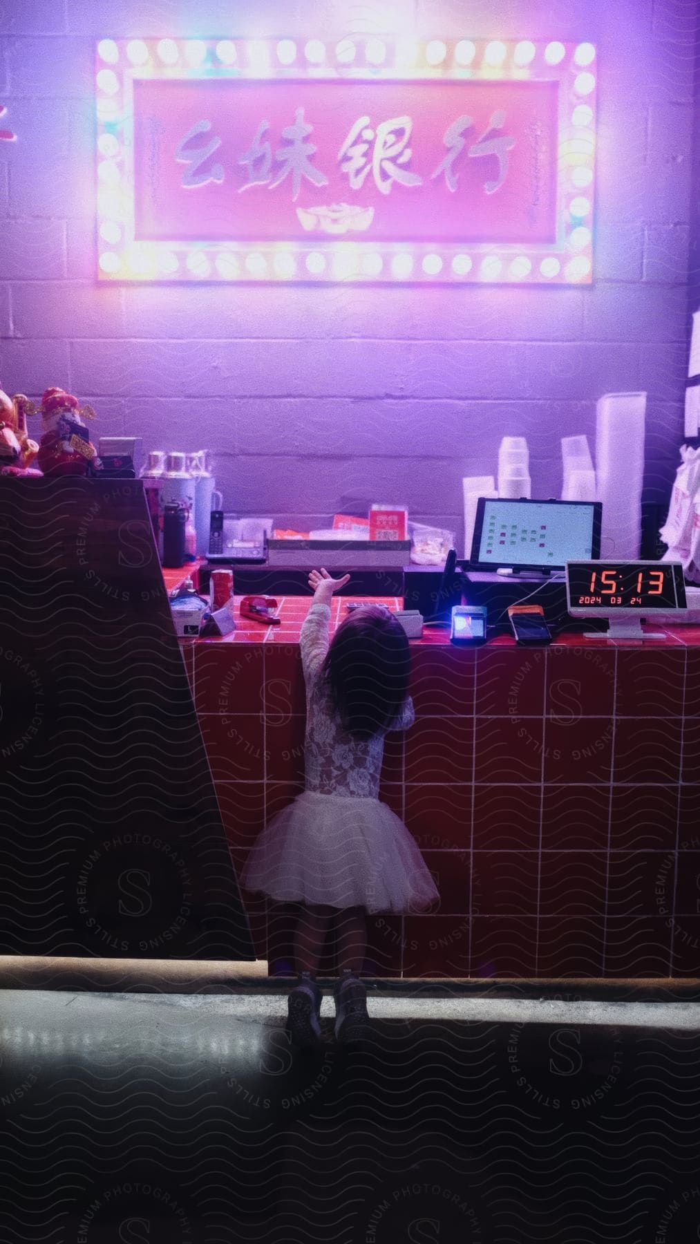 A little girl in a dress is reaching to grab something from a red counter with some electronics on it.