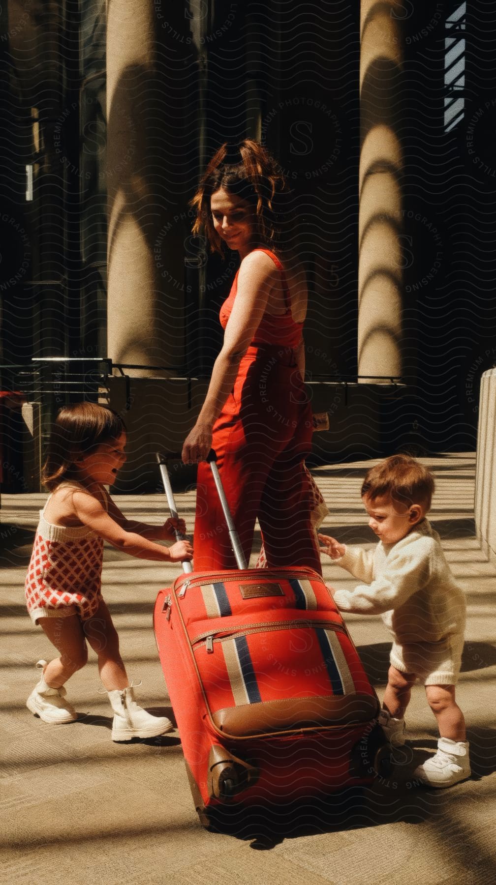 A woman in a red dress rolls a red suitcase on the sidewalk as her two kids play with it while walking together on the street.