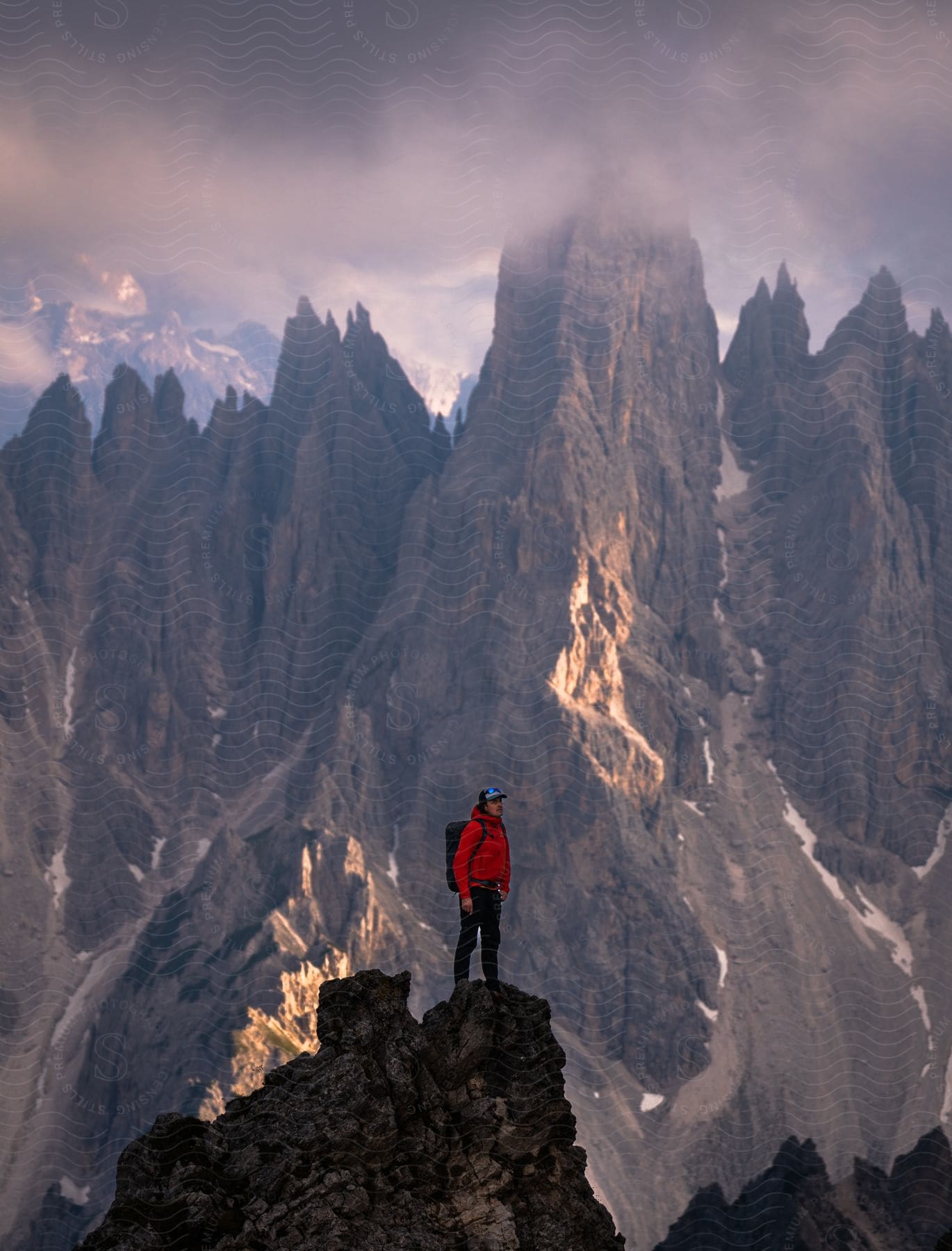 A person in a red coat is standing on a rocky peak in an impressive view of sharp and pointed rocky mountain peaks under a cloudy sky.