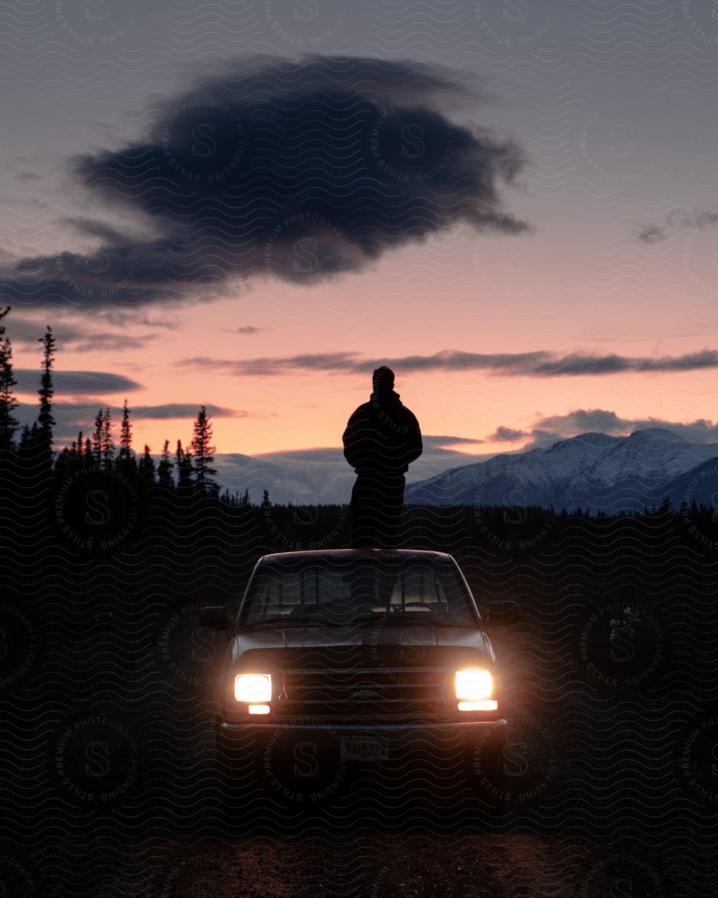 An adult stands on the back of a pickup truck and watches the sun set over a mountain range.