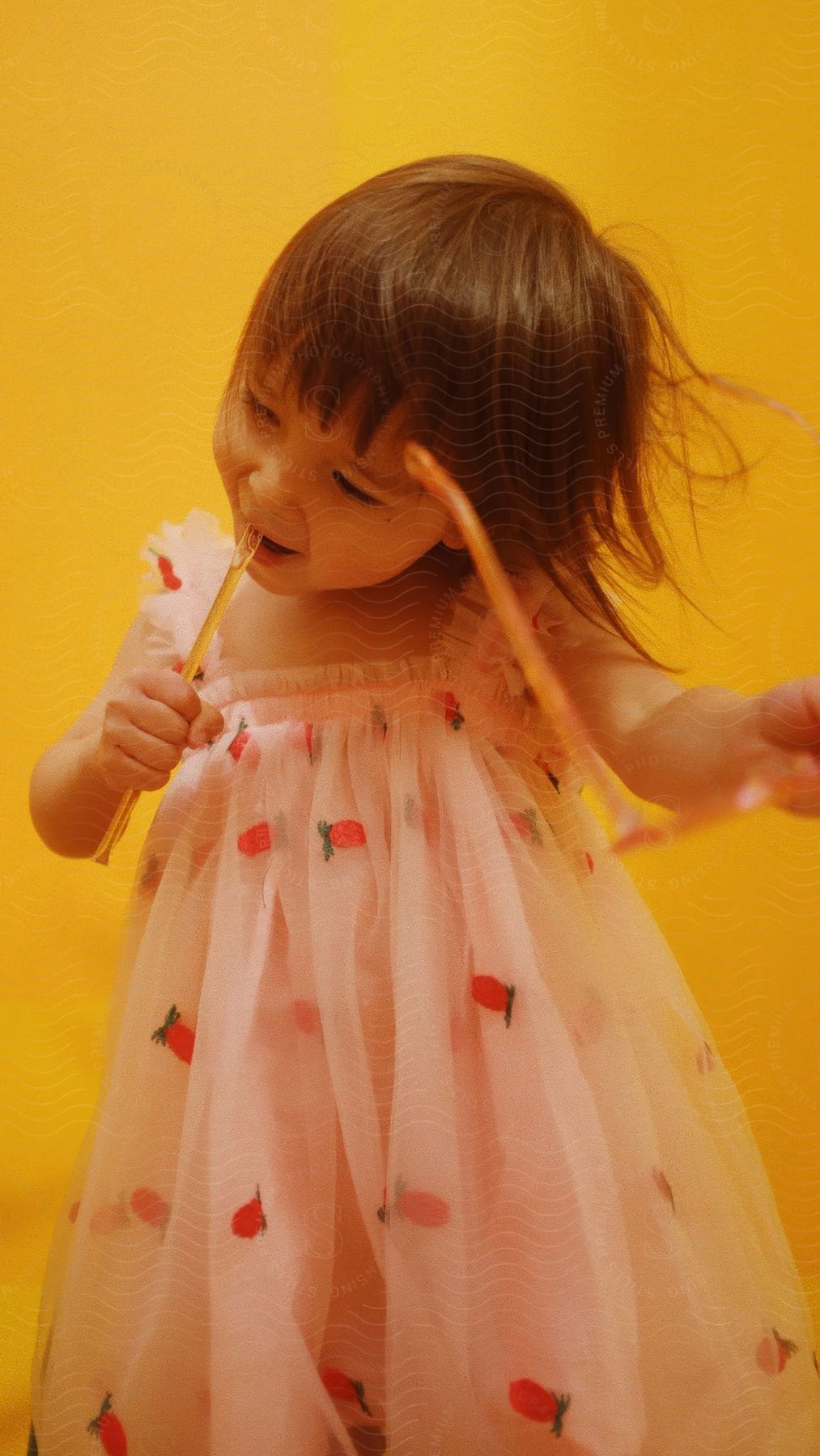 A child holds a pair of yellow sunglasses with a broken handle, placing the broken piece in her mouth as she plays with it. The room has a yellow background.