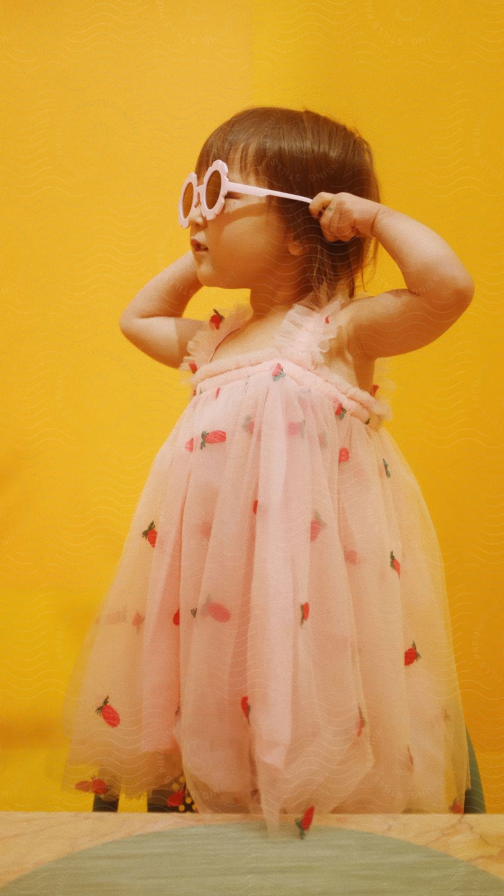 Little girl in a strawberry print dress with her hands in flowered sunglasses on a yellow background.