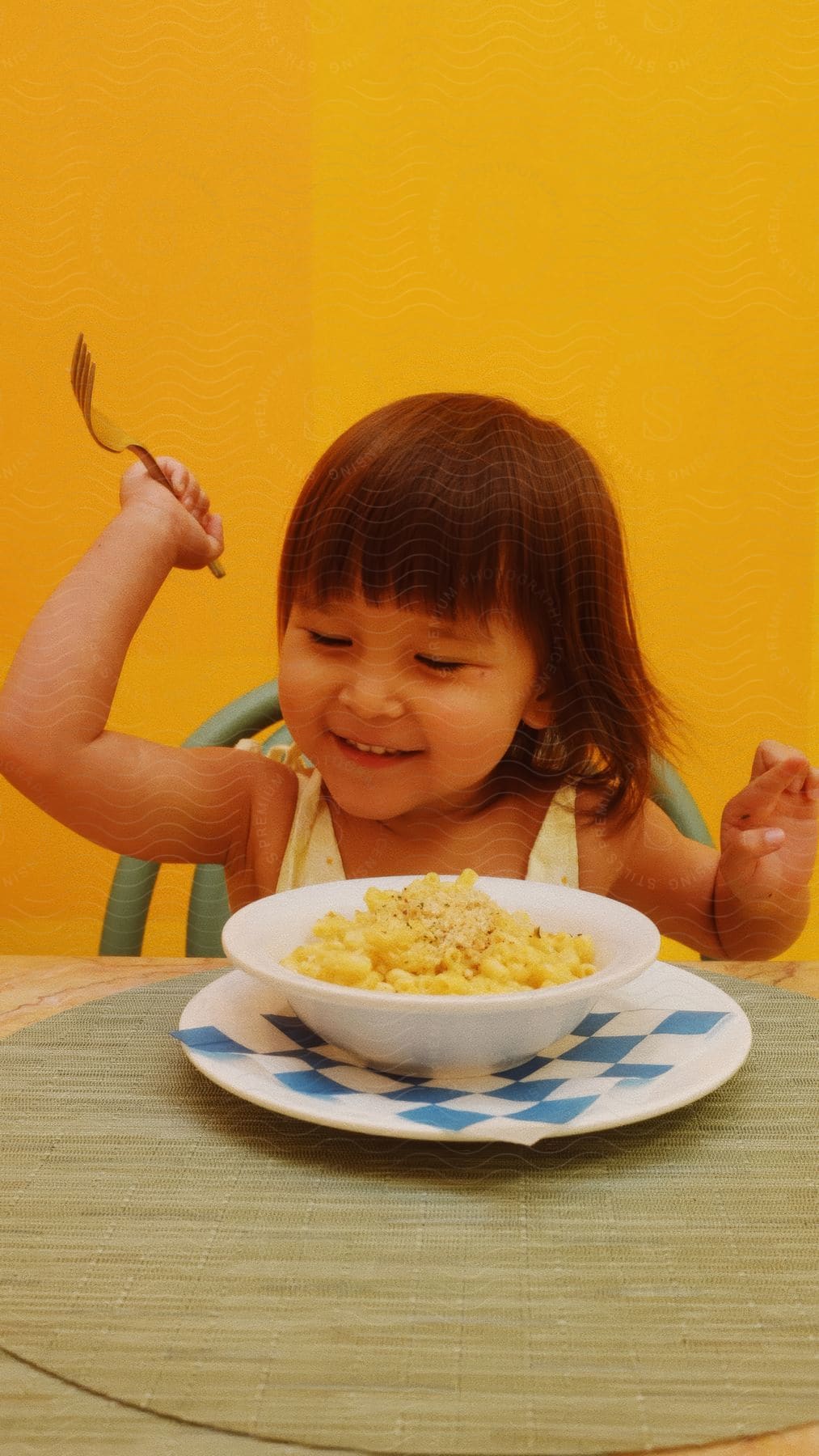 Young child smiling while holding a fork over a bowl of macaroni and cheese, seated at a table against a yellow background.