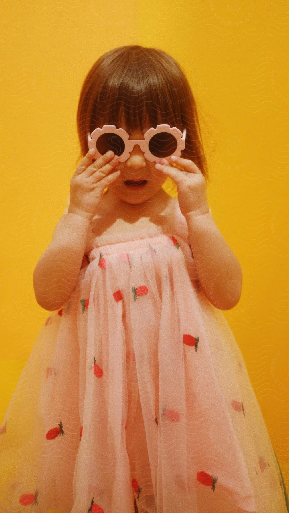 Girl with her hands in flower-shaped sunglasses against a vibrant yellow background and wearing a pink dress with strawberry prints.