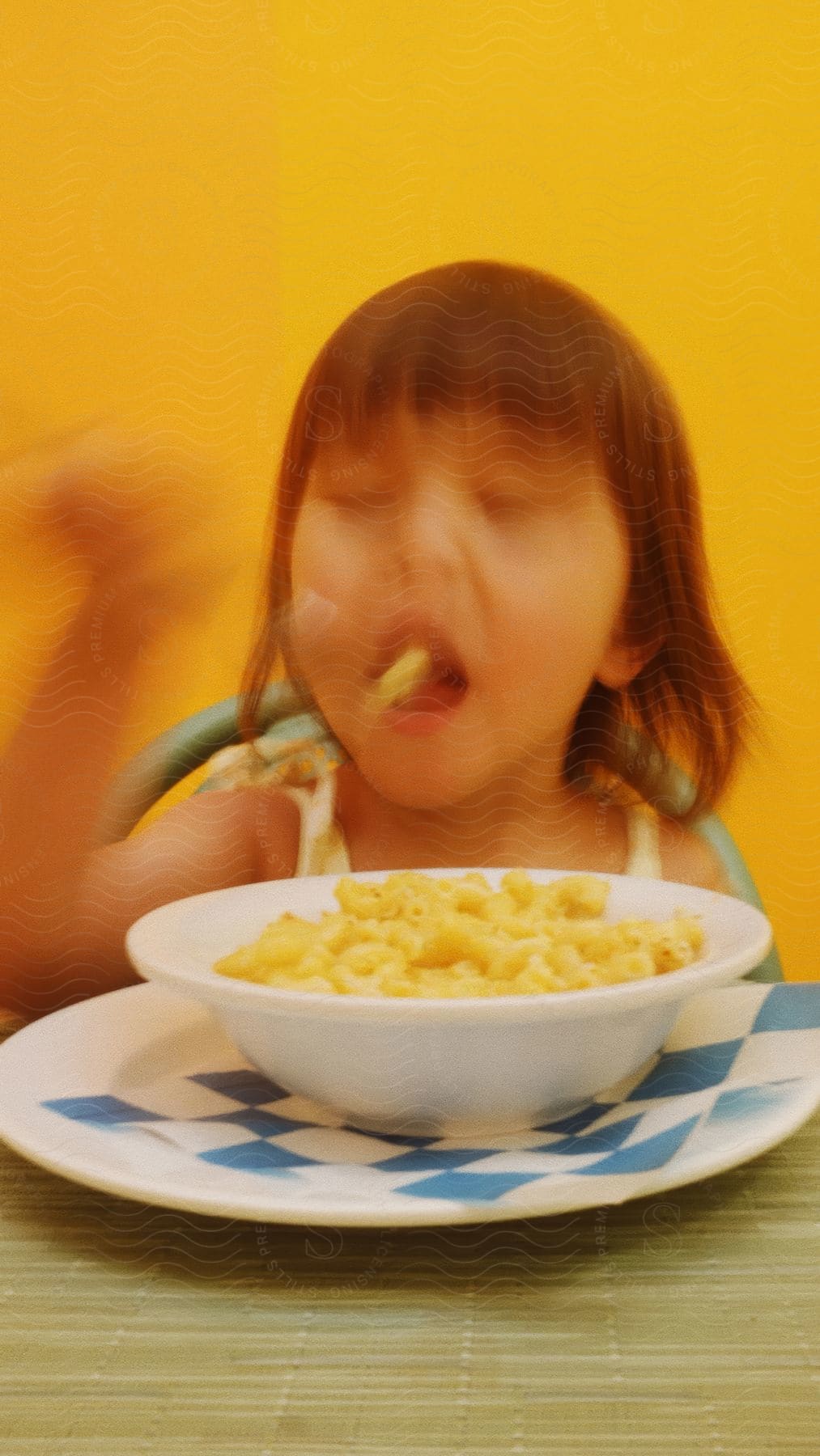 A young Asian girl tosses a piece of Mac and Cheese into her mouth from a bowl in front of her.