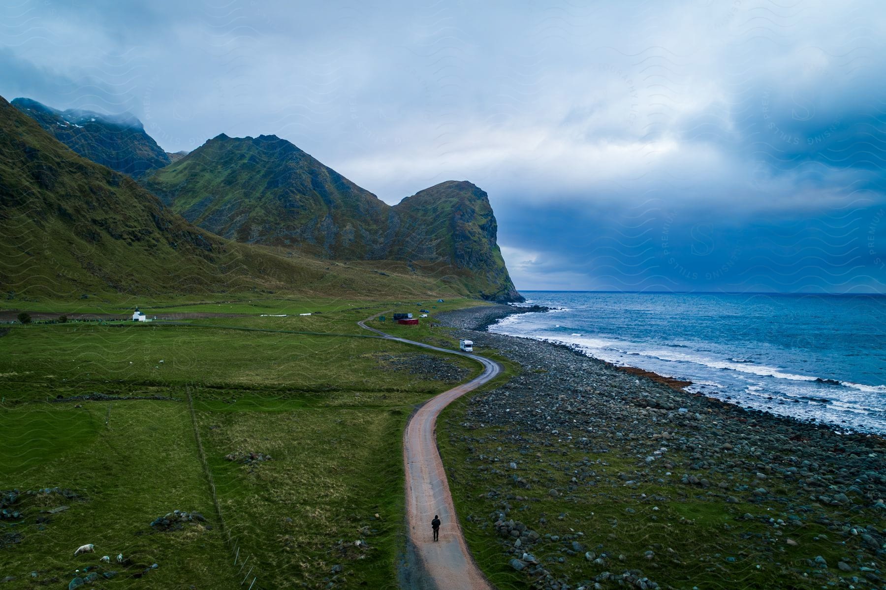 Person walking on a road along the coast as waves roll into shore with mountains in the distance under a cloudy sky