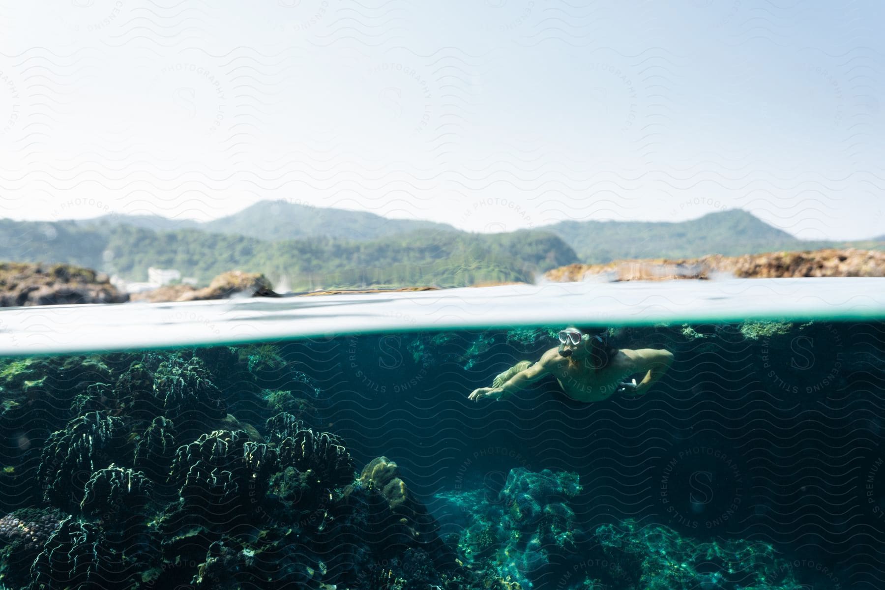 A male is swimming underwater while wearing a snorkle mask in the sea surrounded by mountains.