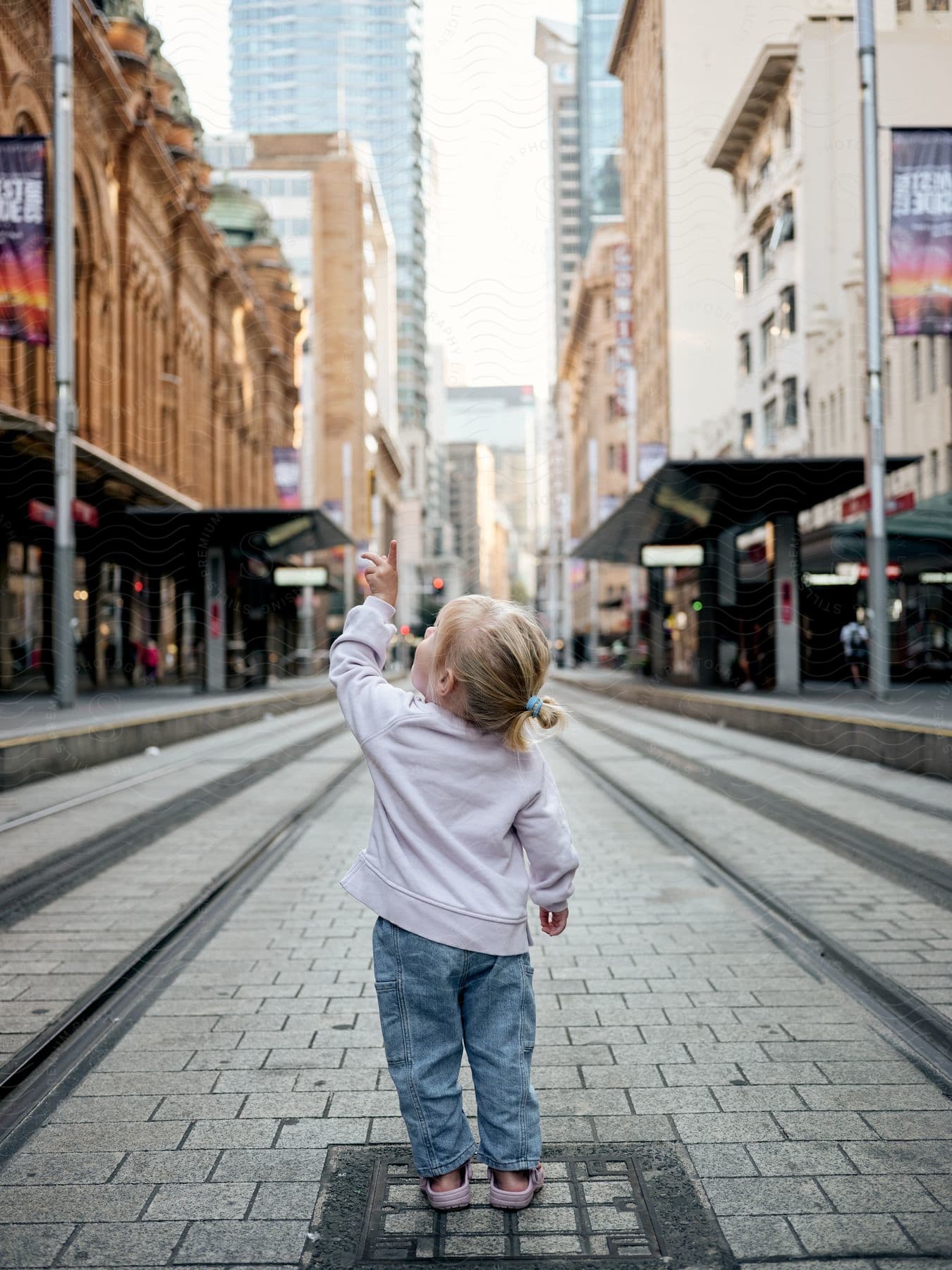 A child stands in the middle of the downtown street during the day, pointing at a building.