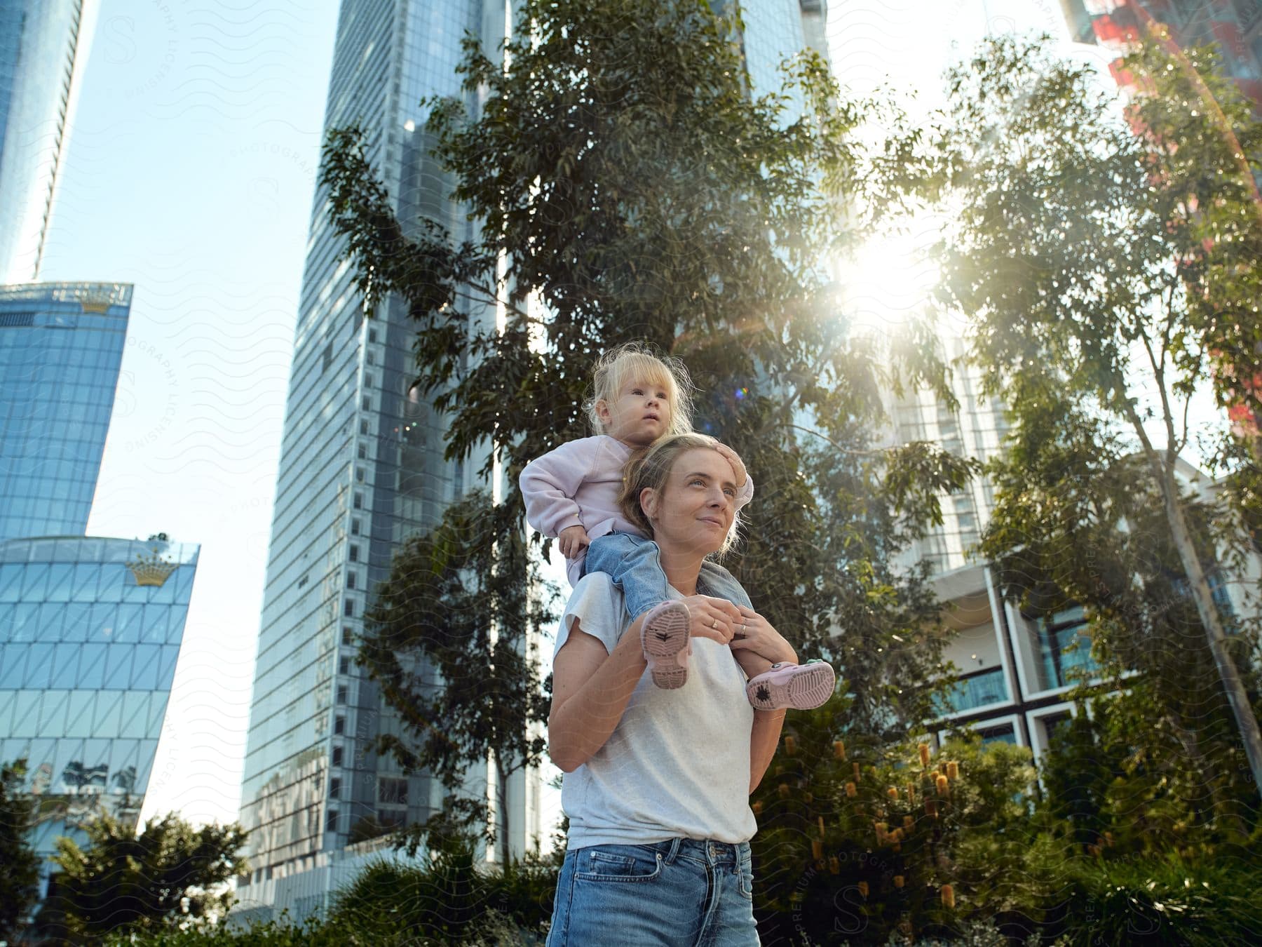 Mother carrying her little daughter on her shoulders while walking in an urban setting with modern skyscrapers.