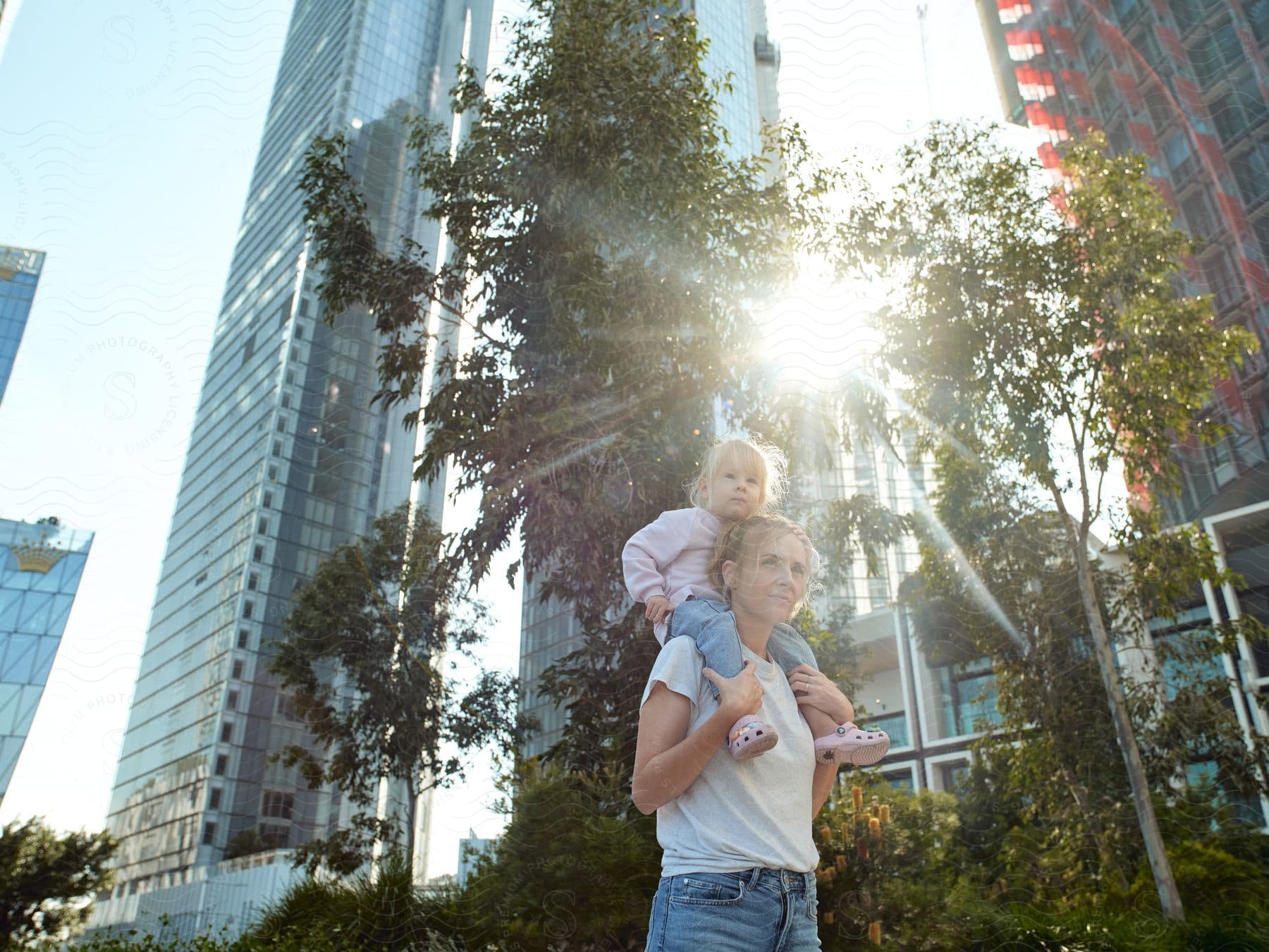 Mother carrying her daughter on her shoulders as they walk through a metropolis with skyscrapers and greenery in the sunlight.