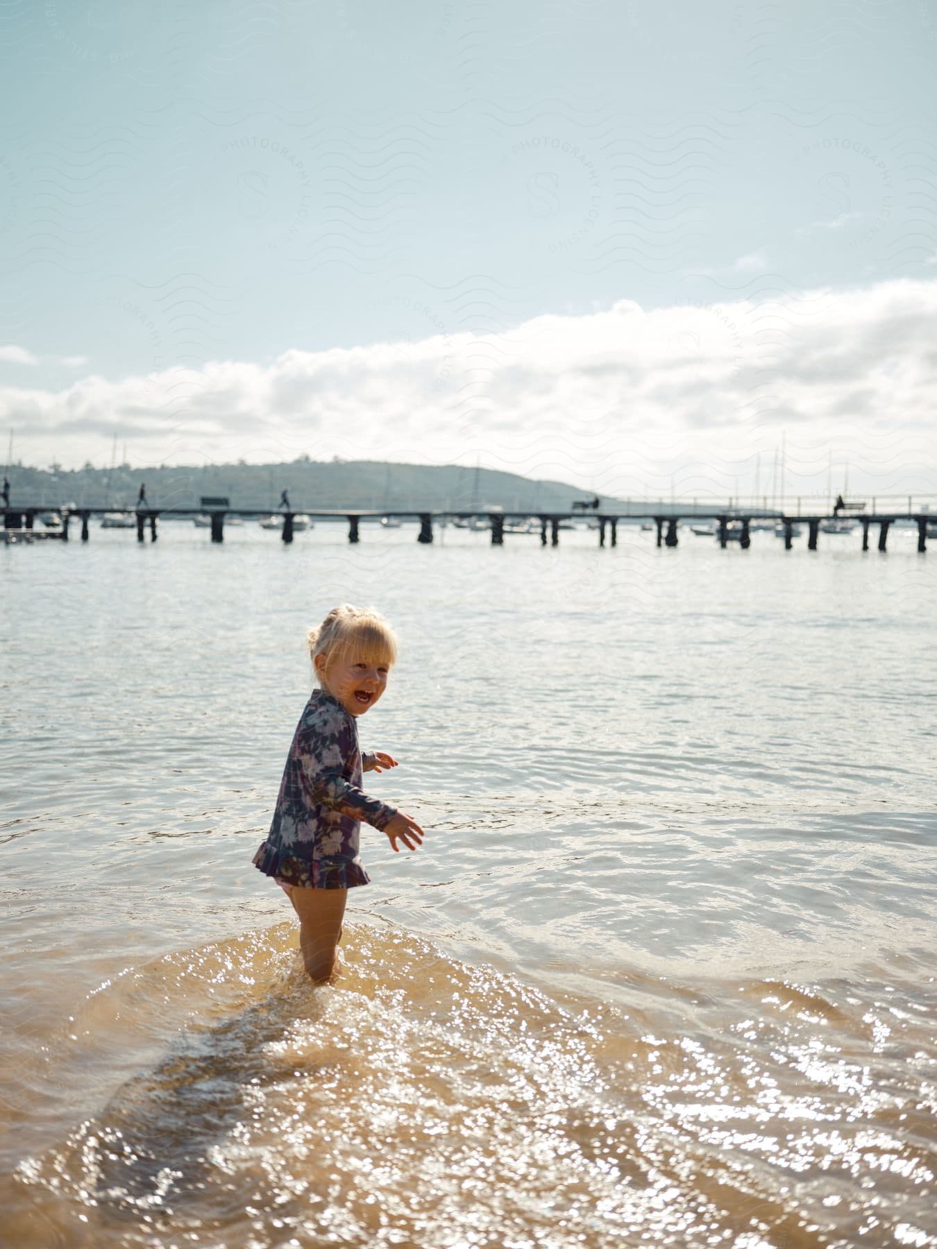 Young girl wades into water across from marina and laughs.