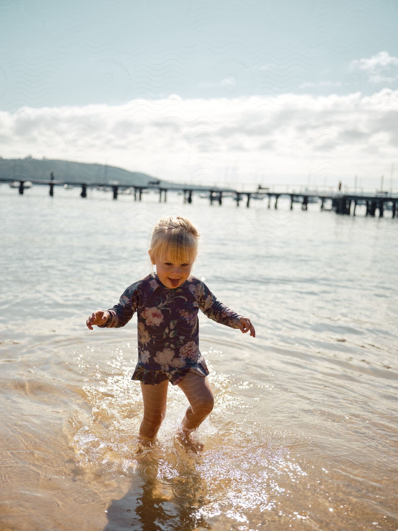 A young girl wading in the water smiling and splashing with a pier behind her