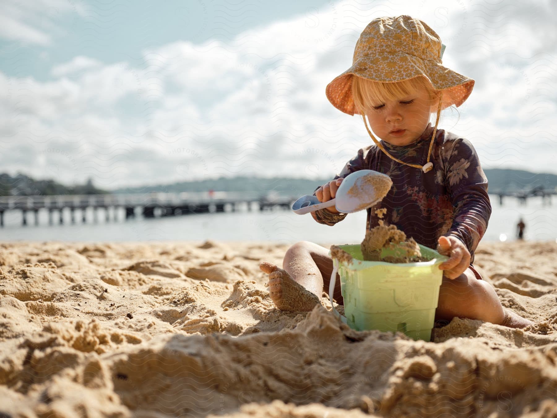 Toddler with a sun hat playing with sand and a bucket on the beach.