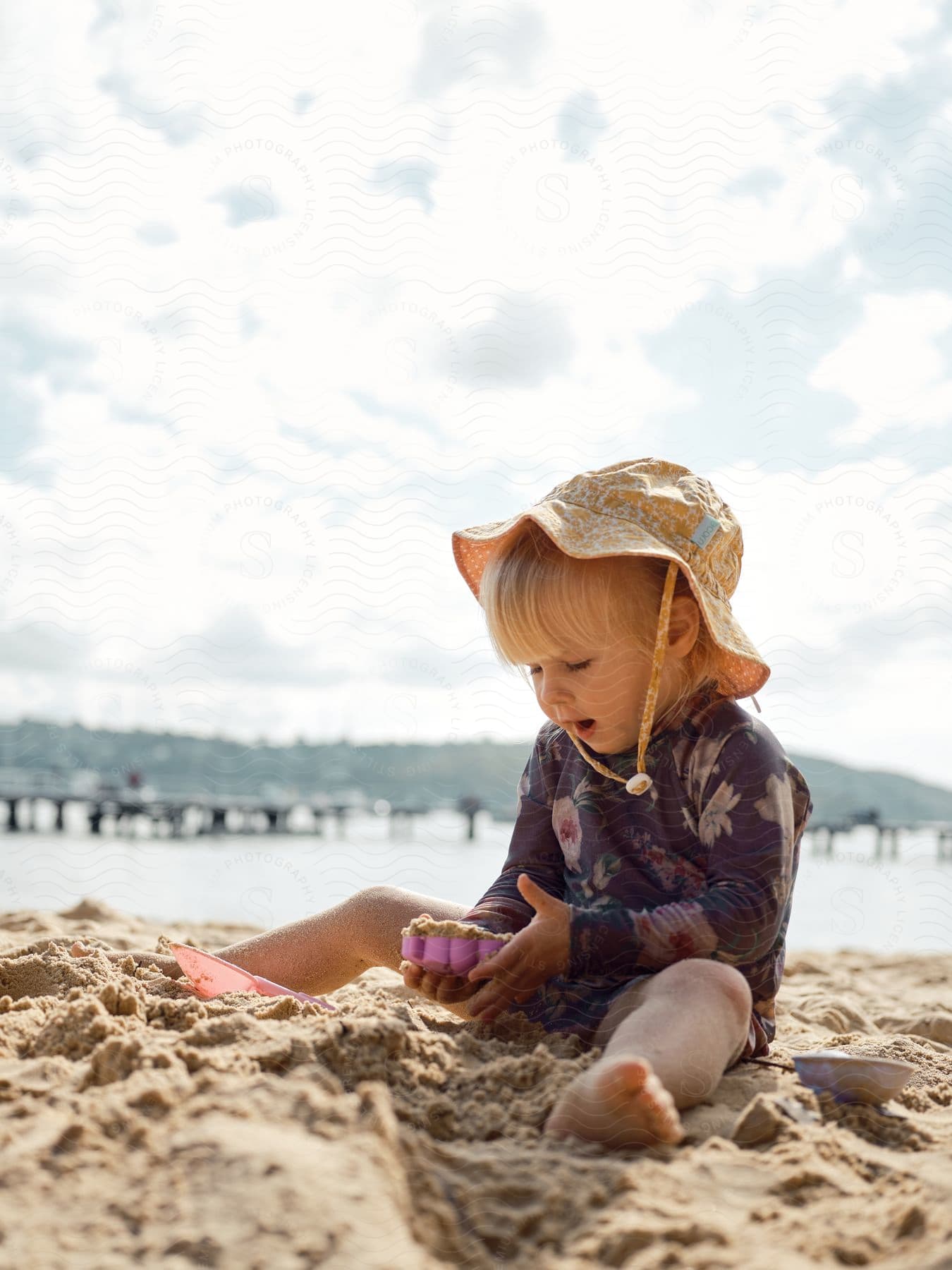 A toddler sits on the beach and plays in the sand on a partly cloudy day.