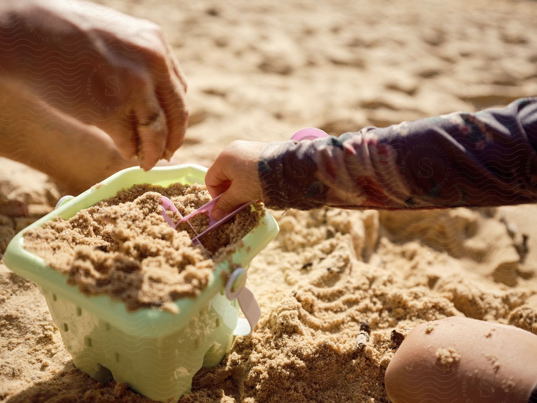 Two people are filling a small green bucket with sand using purple shovels.
