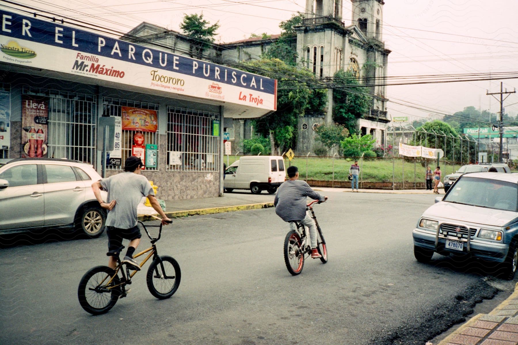 Two males ride bicycles in the street on a cloudy day.