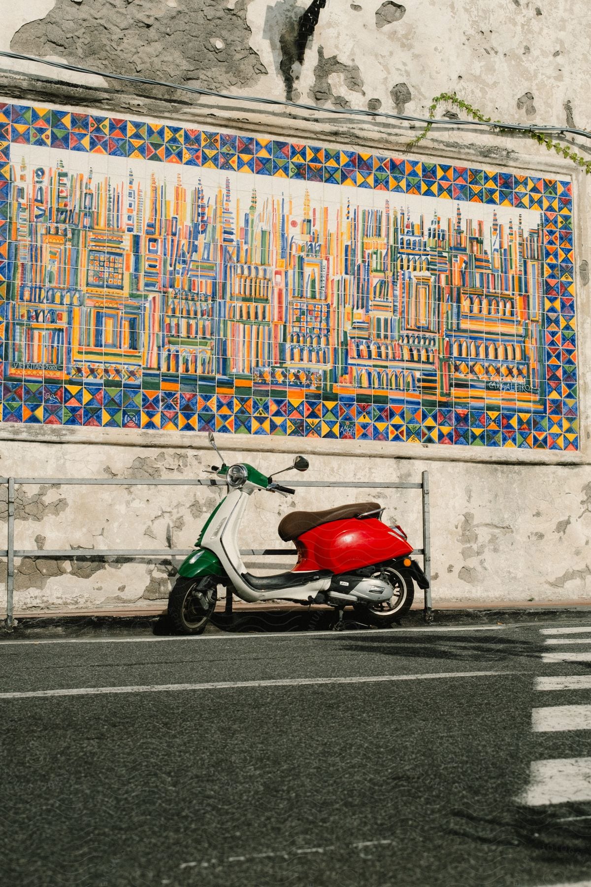 A two wheeled red and white motor scooter on the side of a street next to a wall mural.