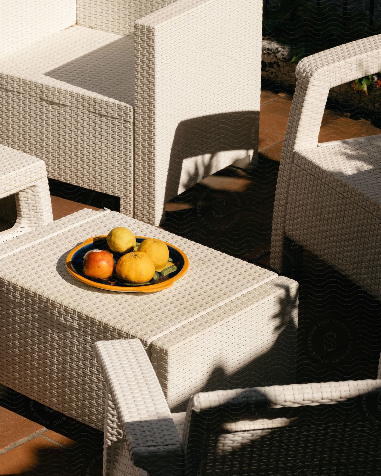 Outdoor white wicker table with a fruit plate in sunlight and chairs around it.