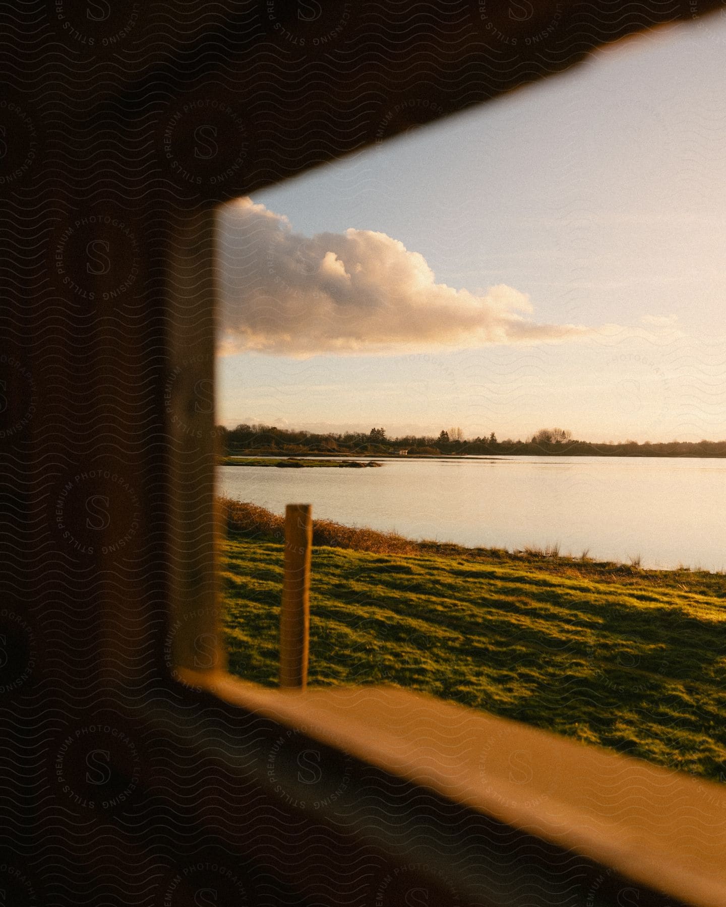 A serene view of a lake and a grassy bank is captured by a wooden window frame, bathed in the warmth of the sunset.